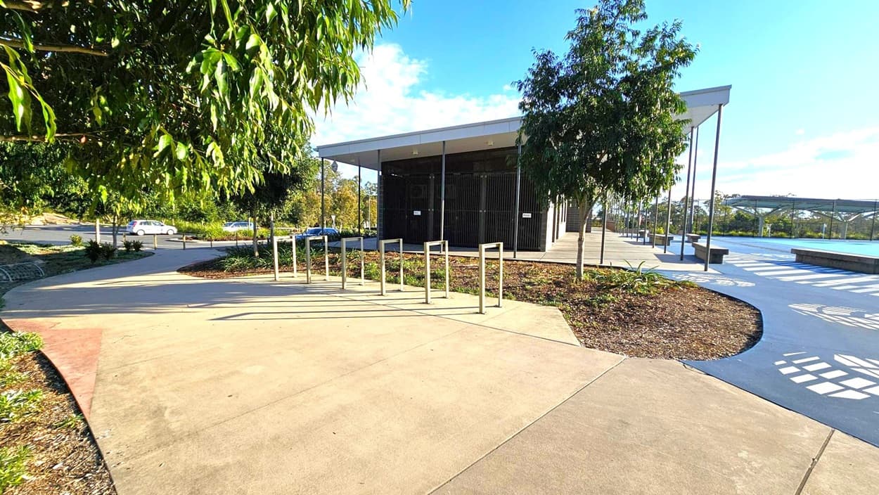 A modern building with a flat roof is situated in the centre, surrounded by a paved area. On the left, there are metal bike racks and a path leading to a car park with a few vehicles visible. Trees and shrubs are planted around the area, providing greenery. On the right, a blue pathway with white patterns runs alongside the building, leading to a covered area with benches. The sky is clear with some clouds.