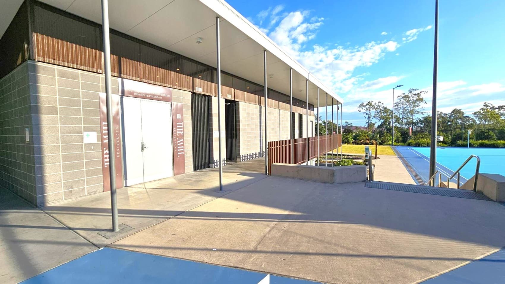 A modern building with a flat roof and a facade of grey bricks and wooden panels on the left. A large door with signage is visible. Several metal poles support the overhanging roof. On the right, there is a swimming pool with clear blue water, bordered by a concrete walkway. In the background, there are trees and a grassy area. The ground is paved with concrete, and the ceiling is flat and white.