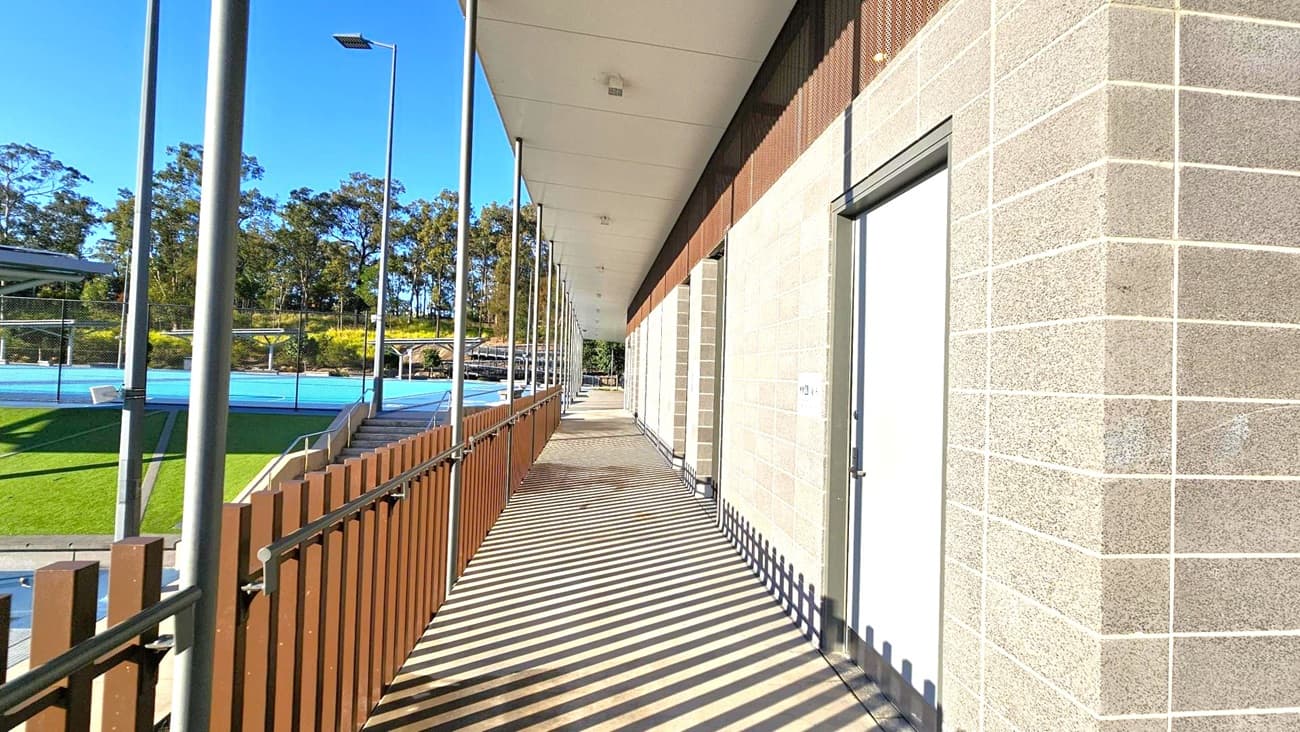 A walkway with a wooden fence on the left and a brick wall on the right. The floor is made of wooden planks, and the ceiling is flat and white. There are tall metal poles supporting the structure. Beyond the fence, there is a blue sports court and green grass. Trees are visible in the background under a clear blue sky.
