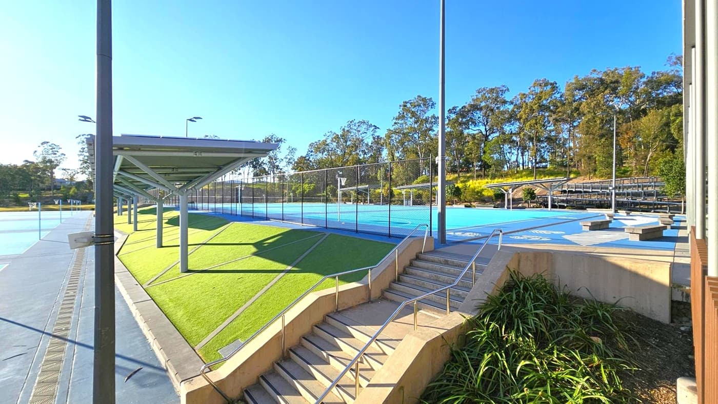 On the left, there is a covered walkway with a metal roof supported by poles, adjacent to a green artificial turf area. In the centre, a blue sports court is enclosed by a chain-link fence. On the right, a set of concrete steps with metal railings leads up to the court, with some plants at the base. In the background, there are trees and a small set of bleachers. The sky is clear and blue.