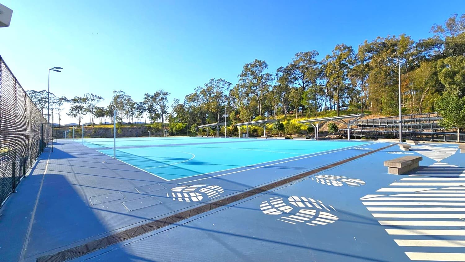 An outdoor sports court with a blue surface and white markings. On the left, a tall black chain-link fence runs alongside the court. In the background, there are trees and a small hill. On the right, there are covered seating areas with metal benches. The foreground features a patterned walkway with white designs and a few concrete benches. The area is well-lit with tall floodlights.