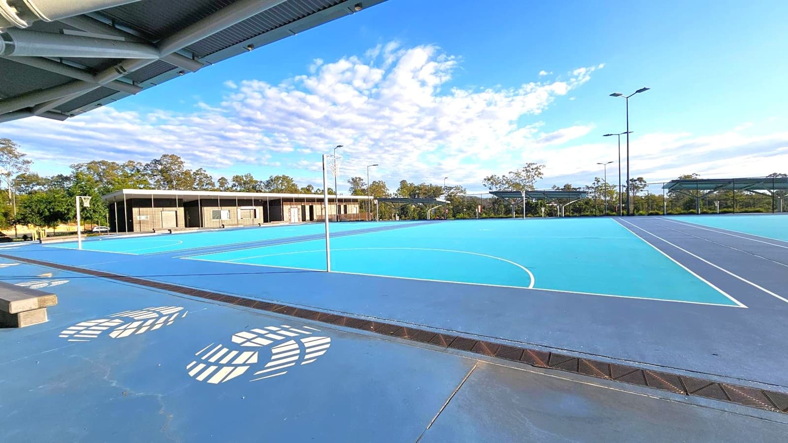 Outdoor sports courts with blue surfaces and white lines, featuring netball hoops. On the left, a covered seating area with a metal roof and large white footprints painted on the ground. In the background, a row of modern, dark-coloured buildings with large windows. Trees and greenery are visible beyond the buildings, and tall floodlights are positioned around the courts.