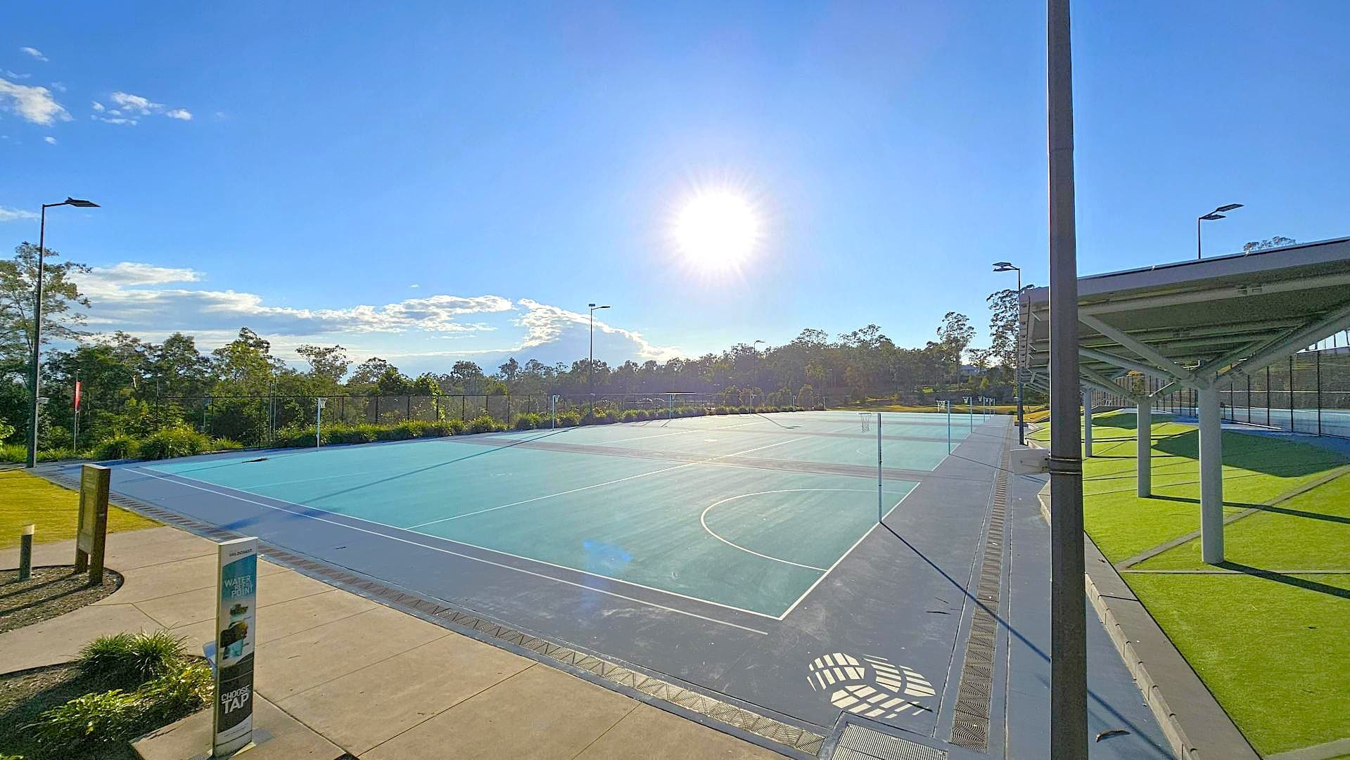 A sports court with a blue surface is in the centre, surrounded by a fence. On the left, there's a concrete path with a water station and some greenery. To the right, a covered area with a green artificial turf is supported by metal poles. The background features trees and a clear sky with the sun shining brightly.