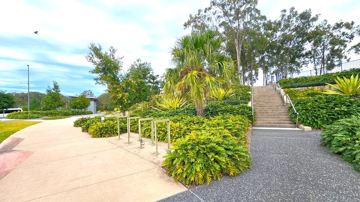 A landscaped outdoor area with a pathway on the left leading to a bike rack. Lush green bushes and small trees line the path. On the right, a set of stairs with metal railings ascends through dense foliage. Tall trees are visible in the background. The ground is a mix of smooth concrete and textured surfaces.