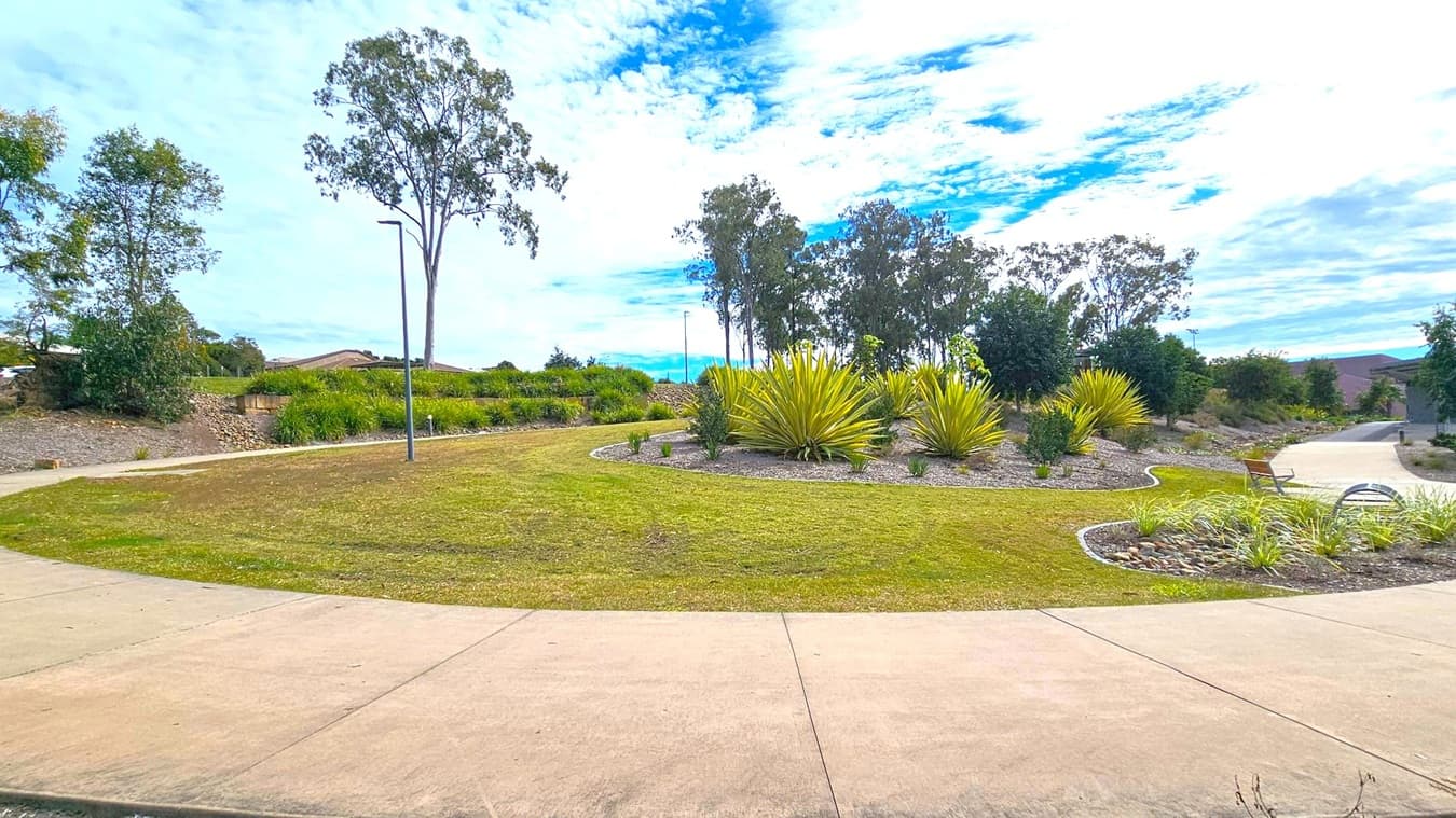A landscaped park area with a curved concrete path in the foreground. The middle features a circular garden bed with spiky green plants and small shrubs, surrounded by grass. To the right, a bench is placed near the path. Tall trees are scattered throughout the background, with a few streetlights visible. The sky is partly cloudy, casting a soft light over the scene.