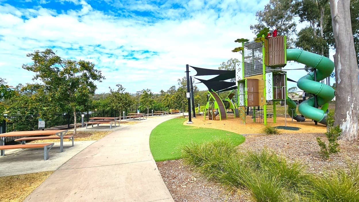 A park scene with a paved path running through the centre. On the left, there are several picnic tables with benches under small trees. On the right, a playground features a tall structure with a green spiral slide and climbing elements. The ground is covered with mulch and artificial grass. In the background, there are more trees and a fence. The sky is partly cloudy.