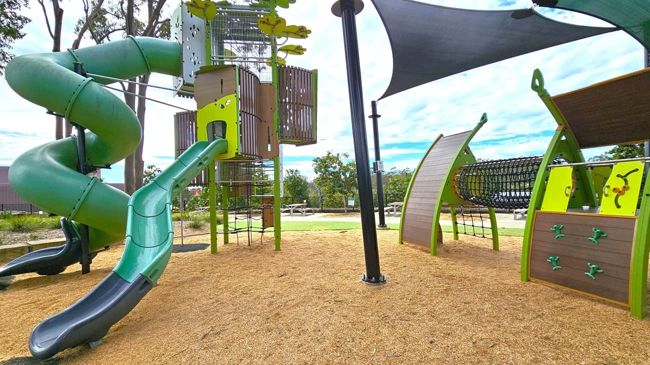A playground with a large green spiral slide on the left, connected to a tall climbing structure. In the centre, there's a vertical climbing net and a small slide. On the right, a climbing wall and a tunnel made of netting are visible. The ground is covered with wood chips, and there are shade sails overhead. In the background, there are trees and picnic tables on a grassy area.
