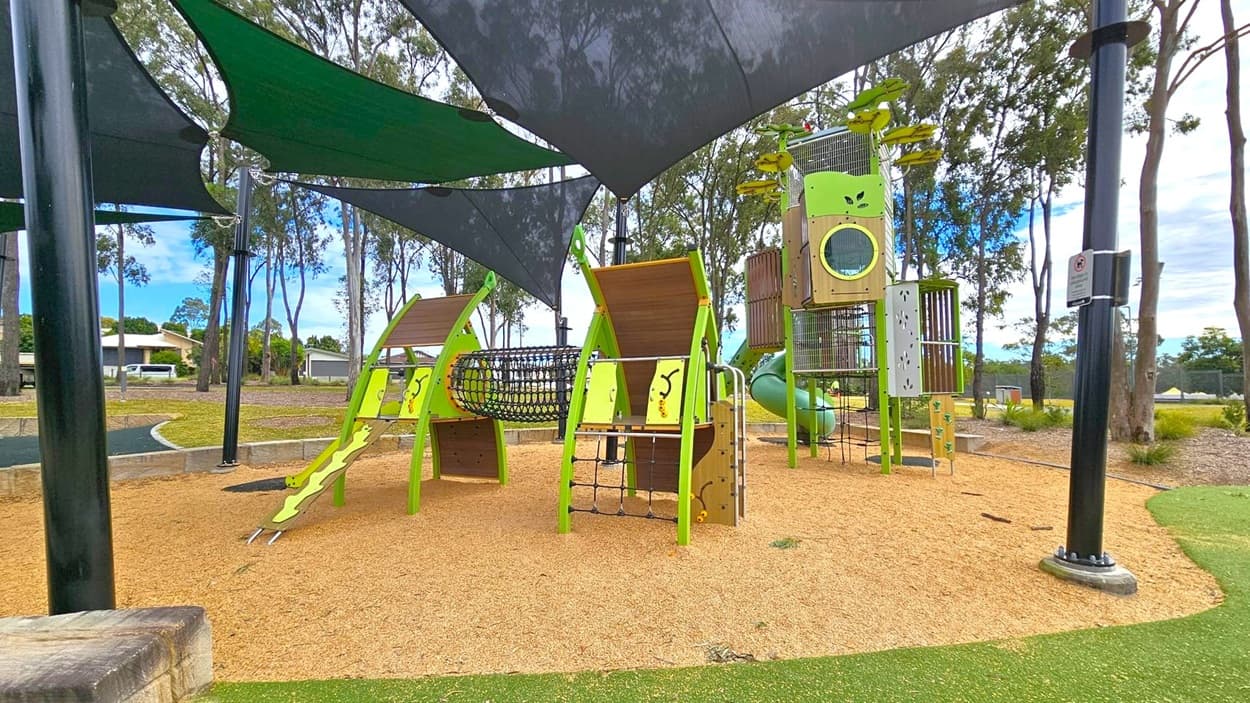 A playground with various climbing structures and slides, primarily in green and brown. The ground is covered with tan mulch, and there are black shade sails overhead. In the background, there are tall trees and a grassy area. A black pole with a sign is visible on the right. The playground equipment includes a net tunnel and a tall tower with a slide.