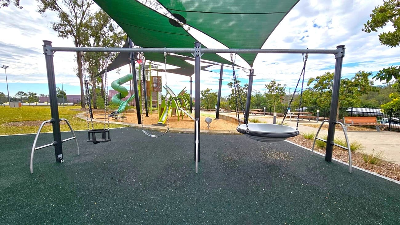 A playground with a swing set in the foreground, featuring a toddler swing and a circular disc swing. The ground is covered with dark rubber matting. In the background, there is a green spiral slide and climbing structures. Green shade sails are overhead, providing cover. To the right, there are benches and a path lined with small plants. Trees and a grassy area are visible in the distance.