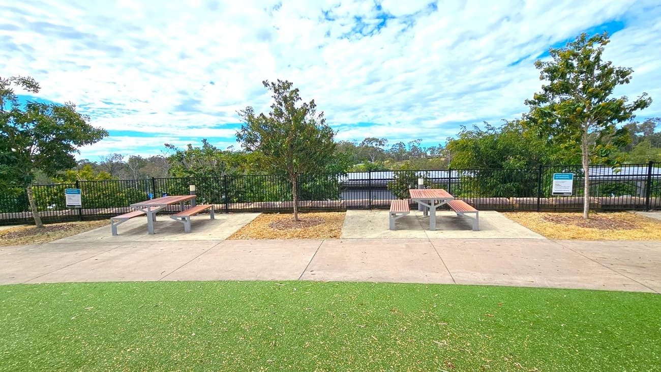 In the foreground, there is a green artificial turf area leading to a concrete path. In the middle, two picnic tables with wooden benches are placed on concrete slabs, separated by small patches of grass with young trees. The background features a black metal fence with dense greenery and trees beyond it. Signs are visible near the tables, and the sky is filled with scattered clouds.
