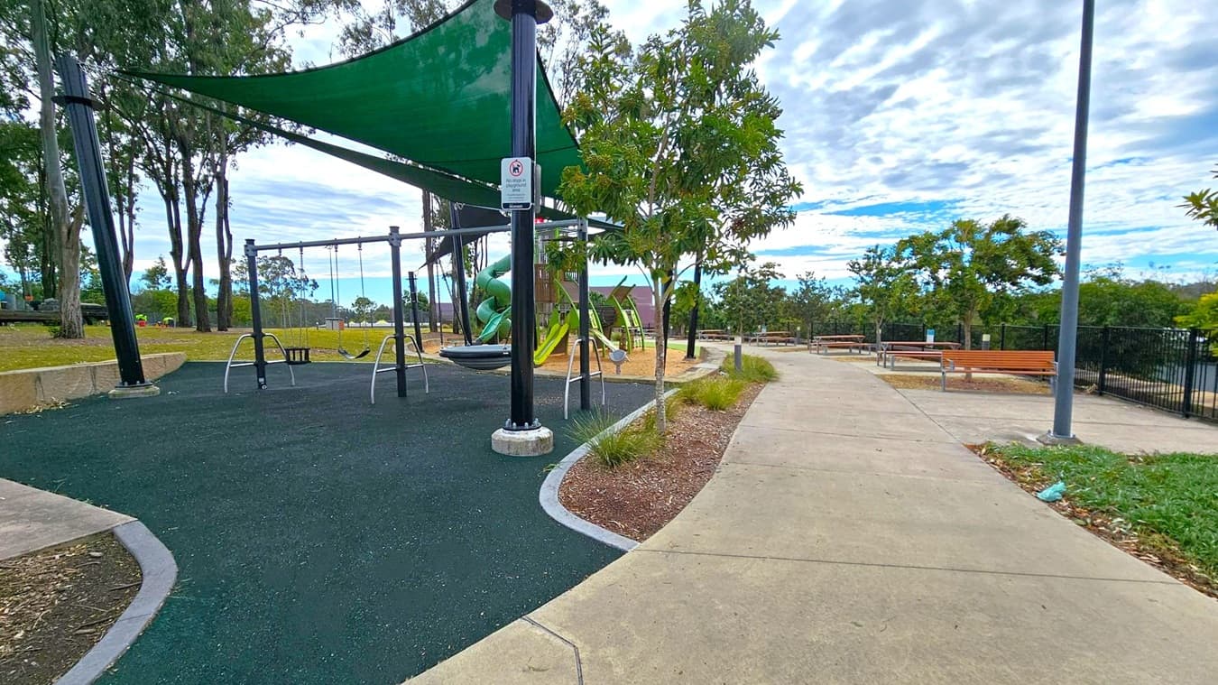 A playground with swings is on the left, covered by a green shade sail. The ground is covered with dark rubber matting. Behind the swings, there's a green spiral slide. Trees and a grassy area are visible in the background. On the right, a concrete path leads past wooden benches and a lamppost. The path is bordered by small plants and a fence is visible further back. The sky is overcast with scattered clouds.