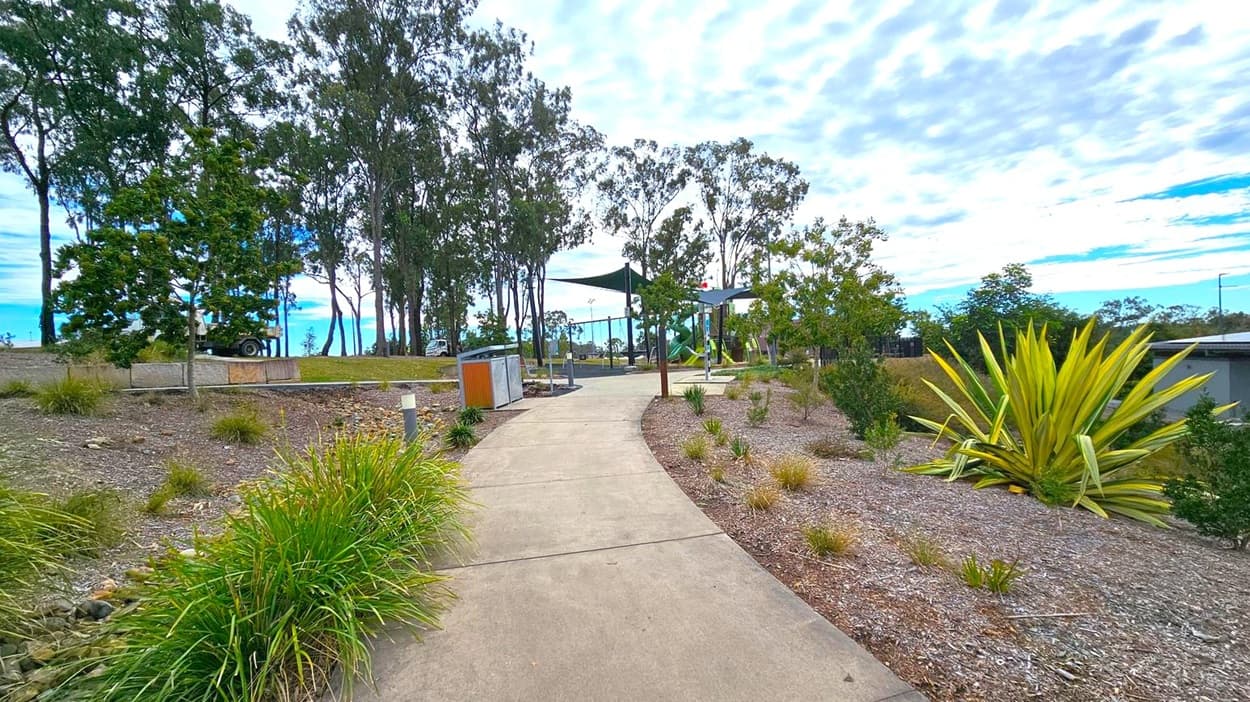 A winding concrete path leads through a landscaped area with various plants and shrubs on both sides. On the left, there are tall trees and a parked vehicle partially visible. To the right, a large spiky plant is prominent among smaller bushes. In the middle distance, a playground with green shade sails and equipment is visible. The ground is covered with mulch and gravel, and the sky is overcast with scattered clouds.