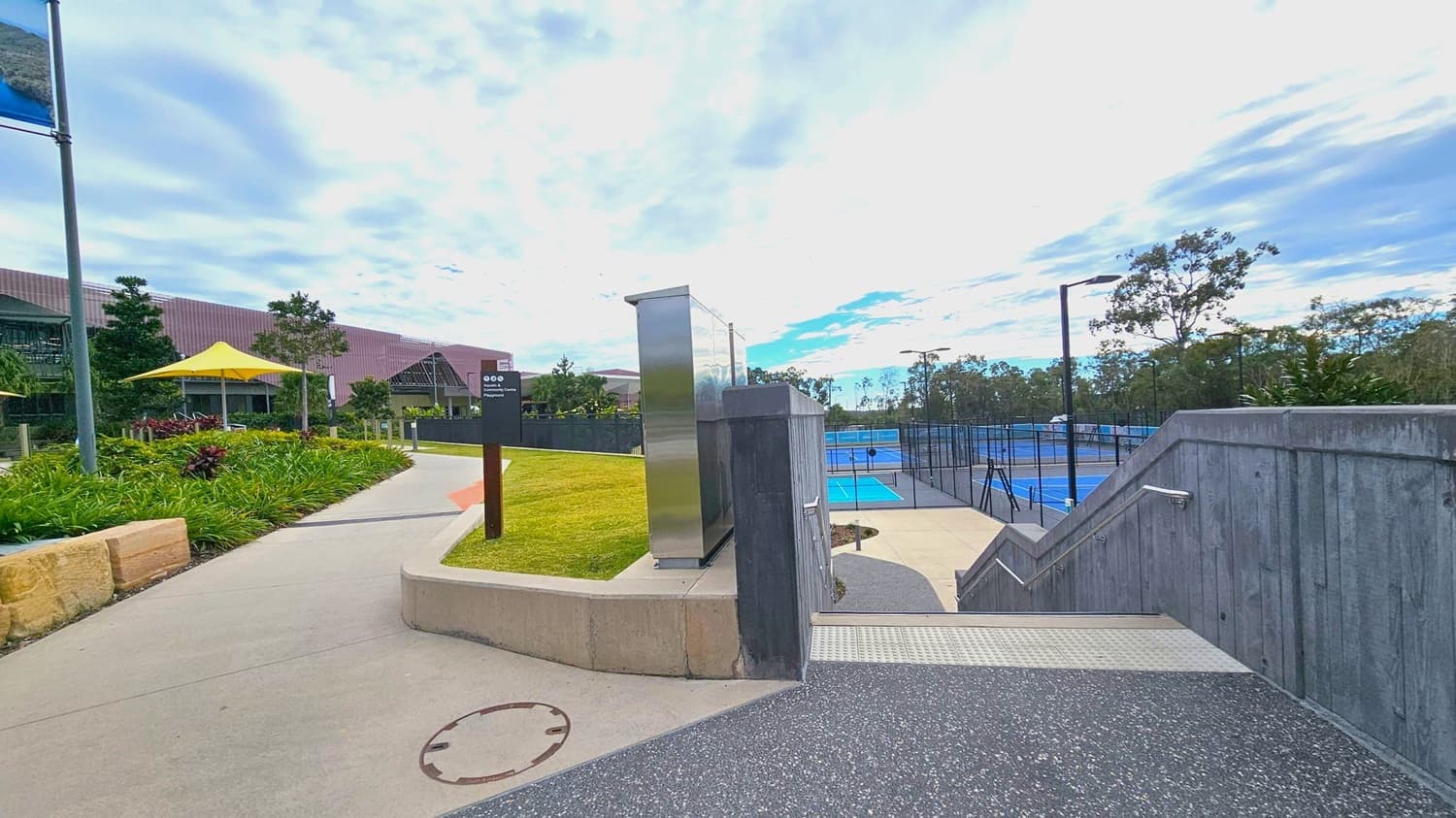 A pathway leads through a landscaped area with green grass and plants on the left. A yellow umbrella provides shade near a building with a sloped roof. On the right, a set of stairs descends to a series of blue tennis courts enclosed by fencing. Metal railings line the stairs, and tall trees are visible in the background. The sky is overcast with patches of blue. A signpost and a metallic structure are near the path.