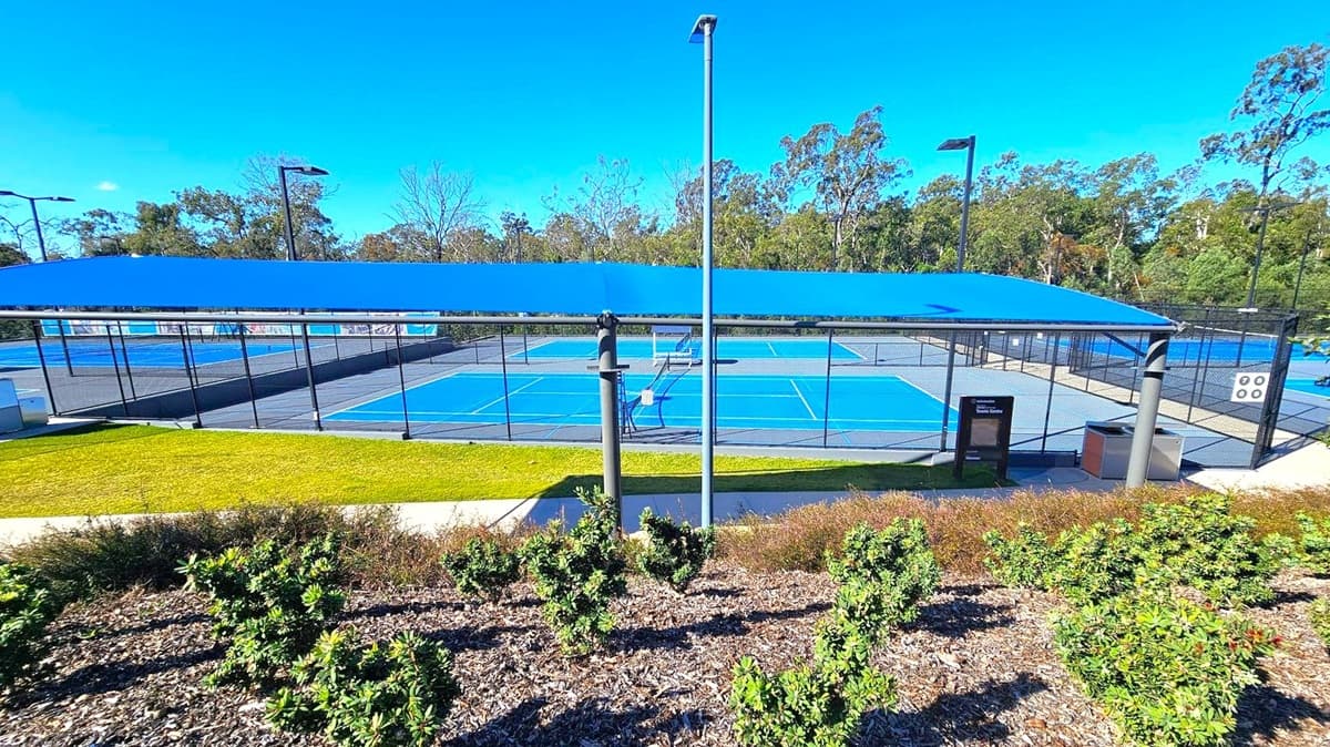 A tennis court complex with multiple blue courts is visible, surrounded by black chain-link fencing. A blue shade structure covers part of the area. In the foreground, there are small green shrubs and a patch of grass. Tall light poles are positioned around the courts. Trees and greenery form the background, with a clear blue sky above. A pathway runs parallel to the courts, and a sign and bin are near the entrance.