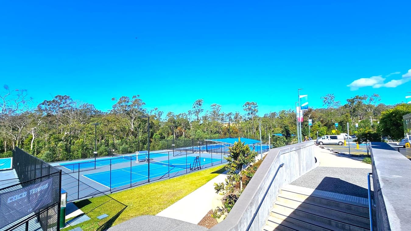 On the left, there are blue tennis courts surrounded by black fencing, with trees in the background. In the middle, a pathway with a small garden area runs alongside the courts. On the right, a concrete ramp with metal railings leads down to a car park where a white van is parked. Banners on poles line the path, and more trees are visible in the distance. The ground is a mix of grass, concrete, and asphalt.