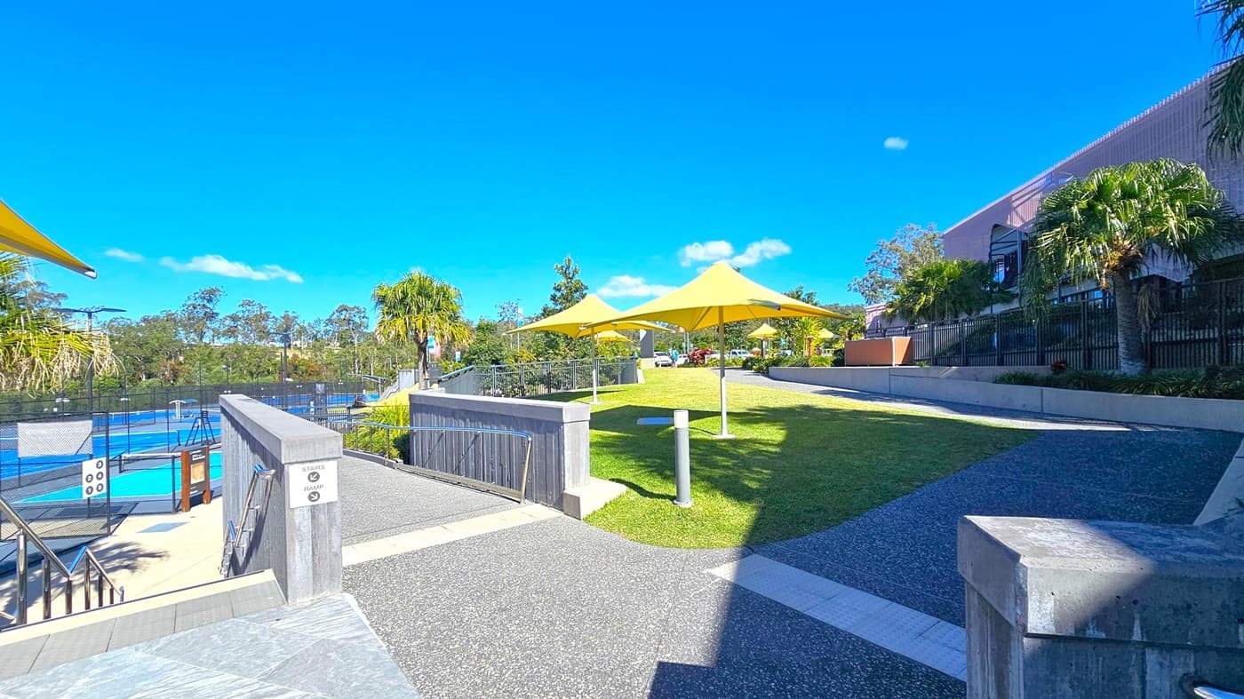 A paved pathway leads through a landscaped area with green grass and several yellow shade structures. On the left, there is a fenced area with blue sports courts. The right side features a building with a modern design and palm trees along the perimeter. The background is filled with trees and a clear blue sky.