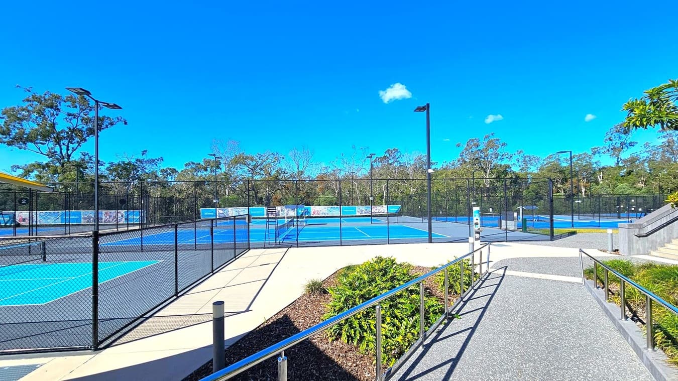 A tennis facility with multiple blue courts surrounded by black chain-link fencing. In the foreground, a pathway with metal railings leads towards the courts. On the right, a set of stairs is visible, bordered by greenery. The background features tall trees and a clear blue sky. A yellow-roofed structure is partially visible on the left.