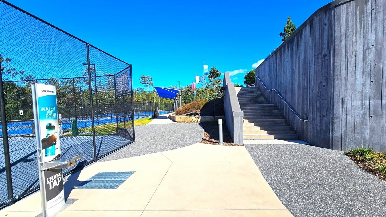 On the left, a chain-link fence encloses blue tennis courts. A water refill station with signage is nearby. In the centre, a paved path leads towards the courts and a grassy area. On the right, a tall concrete wall borders a set of stairs with a metal handrail, leading upwards. Trees and shrubs are visible in the background, and the ground is a mix of concrete and gravel.