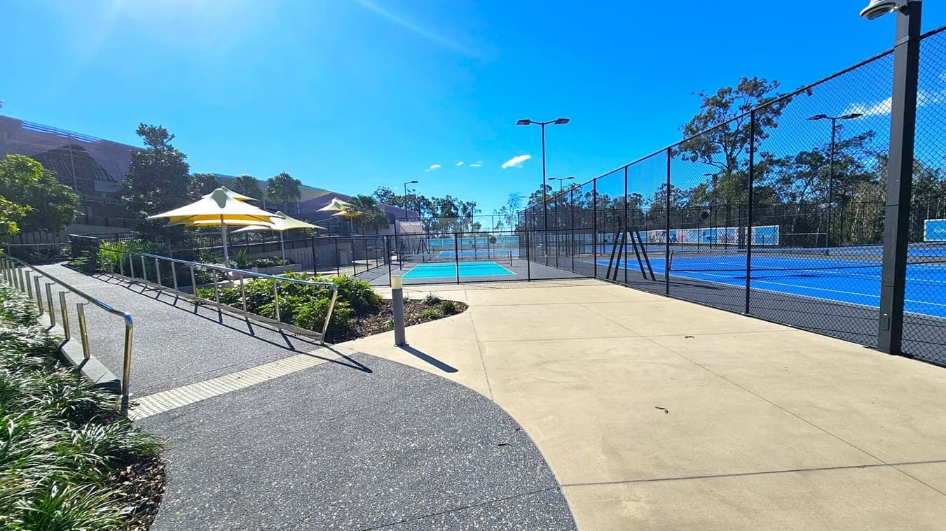 A pathway leads to a fenced tennis court area. On the left, there is a sloped walkway with metal railings and greenery alongside it. Yellow and white umbrellas provide shade over a seating area. On the right, the blue tennis courts are enclosed by a tall chain-link fence. Trees and a building are visible in the background. The ground is a mix of textured and smooth concrete.