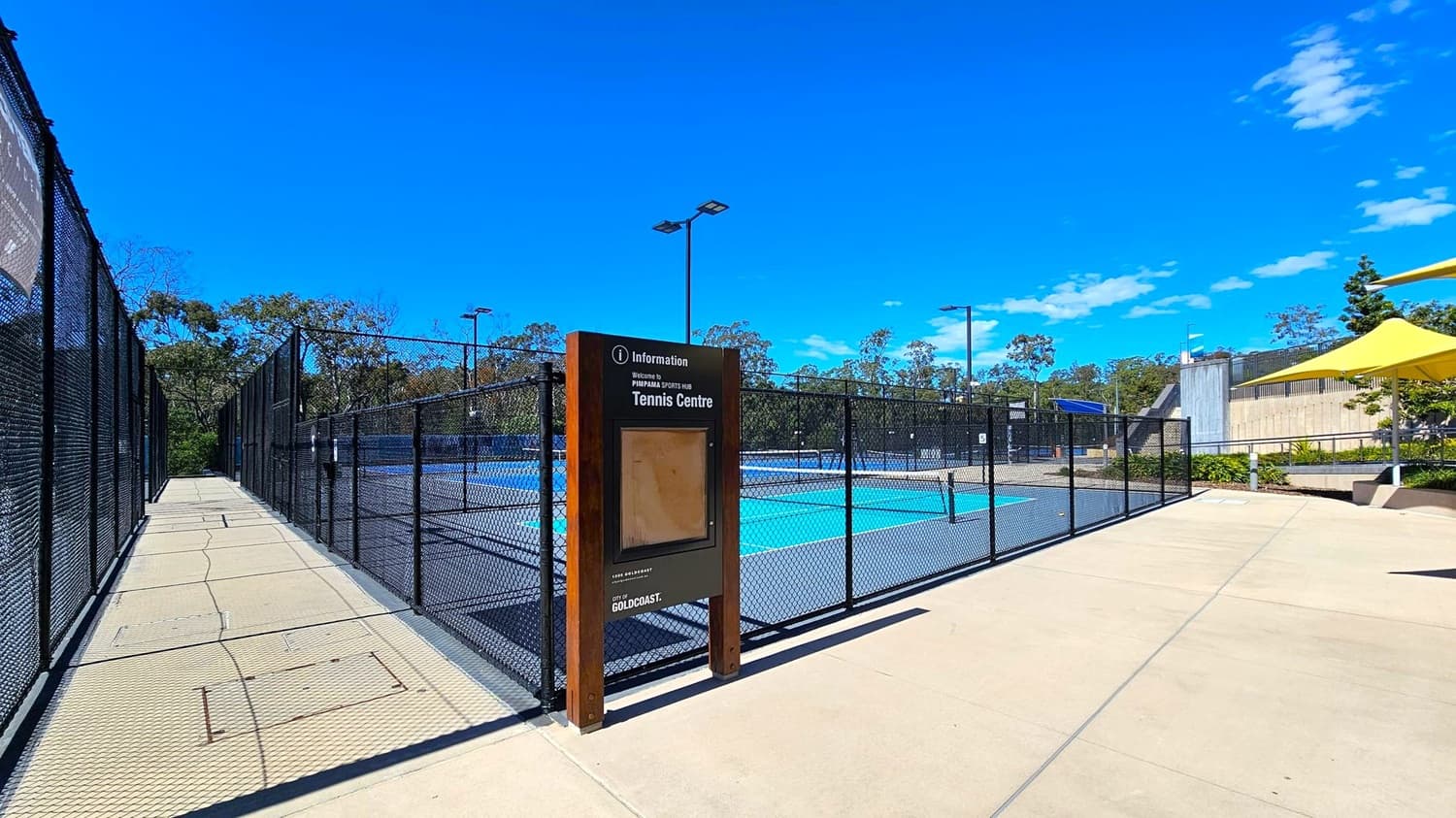 A tennis centre with a fenced court in the middle. On the left, a pathway runs alongside the fence, bordered by trees in the background. In the centre, a signboard provides information about the tennis centre. On the right, a concrete area with a yellow shade structure and some greenery. The ground is paved with light-coloured tiles, and tall lights are visible around the court.