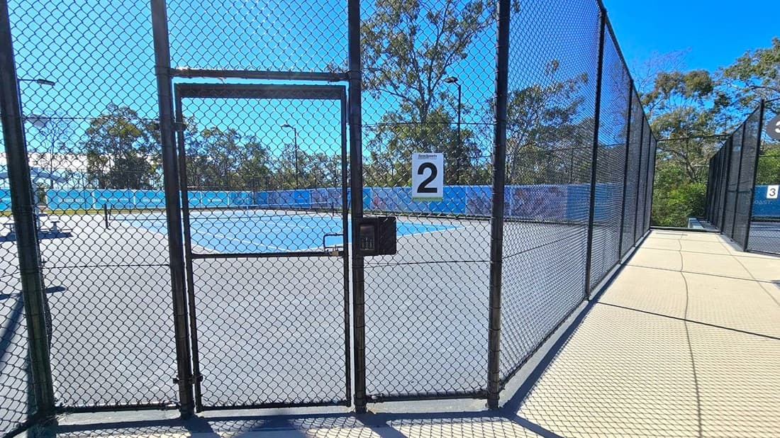 A tennis court enclosed by a chain-link fence with a gate in the centre, marked with the number "2". The court surface is blue and grey. To the right, a pathway runs alongside the fence, leading to another court marked "3". In the background, there are trees and a blue sky. The ground outside the court is a light-coloured concrete.