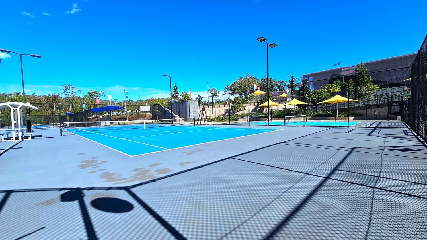 A tennis court with a bright blue playing surface and grey surrounding area. On the left, there is a white bench with a canopy. The court is enclosed by a black chain-link fence. In the background, there are several yellow umbrellas providing shade, and a building partially obscured by trees. Tall floodlights are positioned around the court. The ground is patterned with shadows from the fence and other structures.
