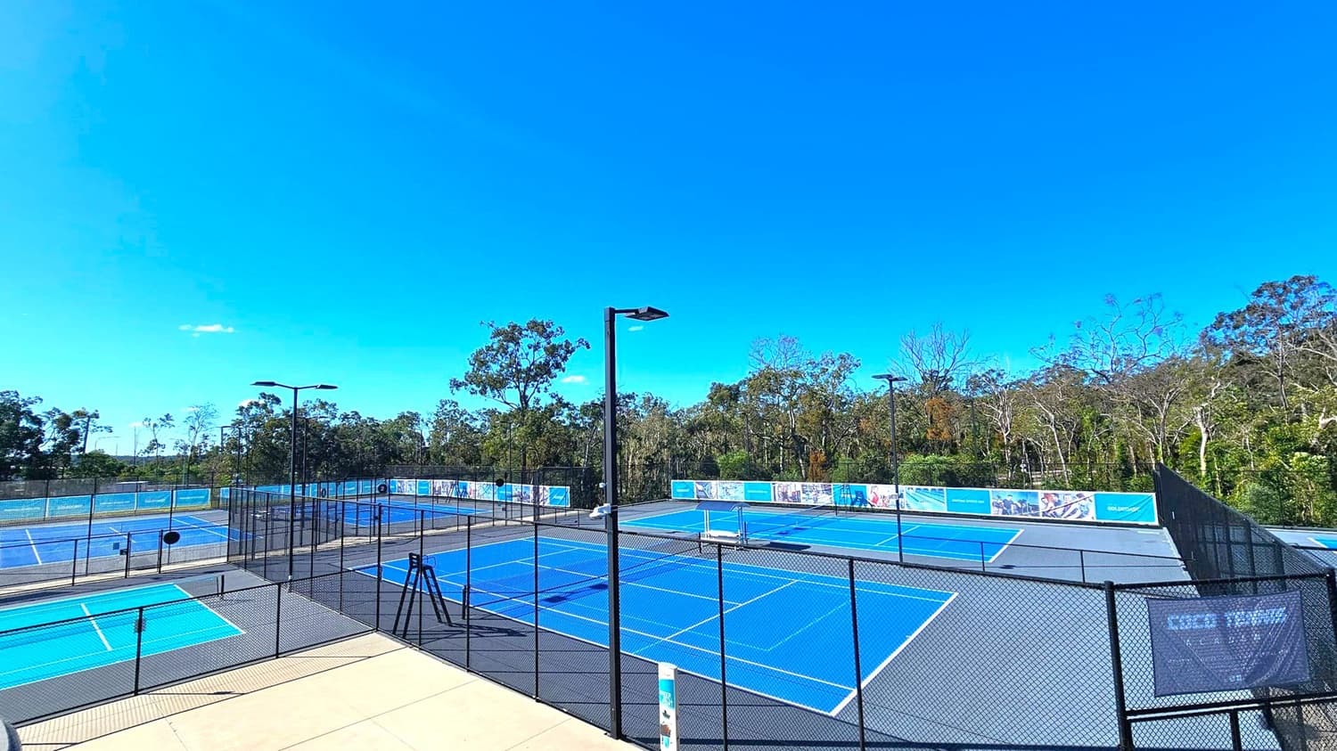 Tennis courts with blue surfaces are surrounded by black chain-link fencing. Several tall floodlights are positioned around the courts. In the background, there is a dense line of trees and vegetation. The courts are empty, and there are banners on the fences. The ground is a mix of concrete and court surfaces.