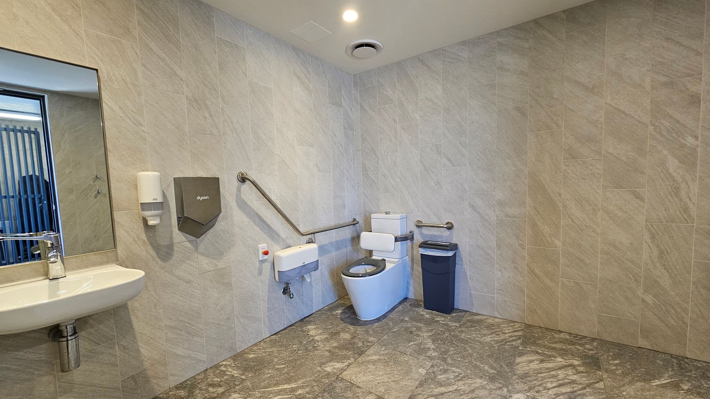 A spacious, accessible bathroom with light-coloured tiled walls and a patterned floor. On the left, a wall-mounted sink with a mirror above it, accompanied by a soap dispenser and a hand dryer. In the centre, a toilet with support rails on both sides and a small bin to the right. The ceiling features a light and a ventilation fan.