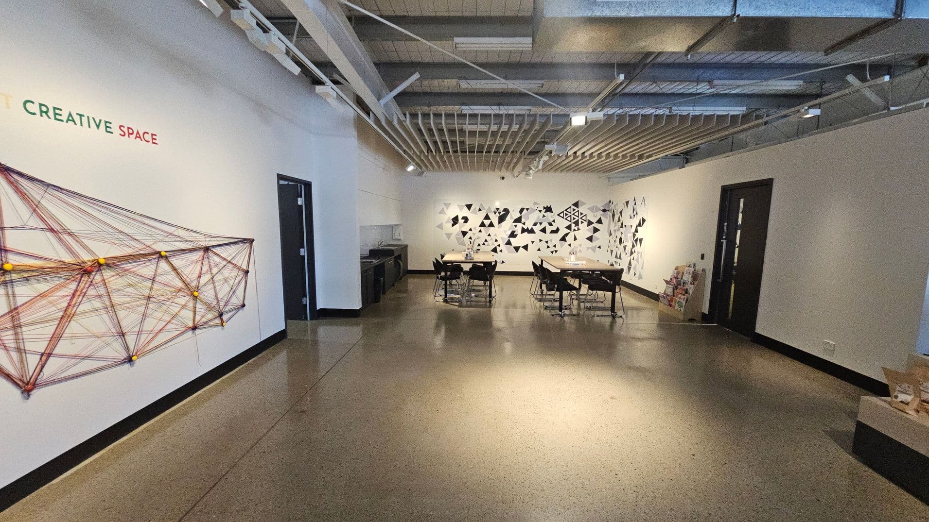 A room with a polished concrete floor and an exposed ceiling with beams and ducts. On the left wall, there's a geometric string art installation with the words "CREATIVE SPACE" above it. In the centre, two tables with chairs are arranged, and the back wall features a pattern of black and white triangles. On the right, there are two black doors and a stack of books or magazines on a low shelf.