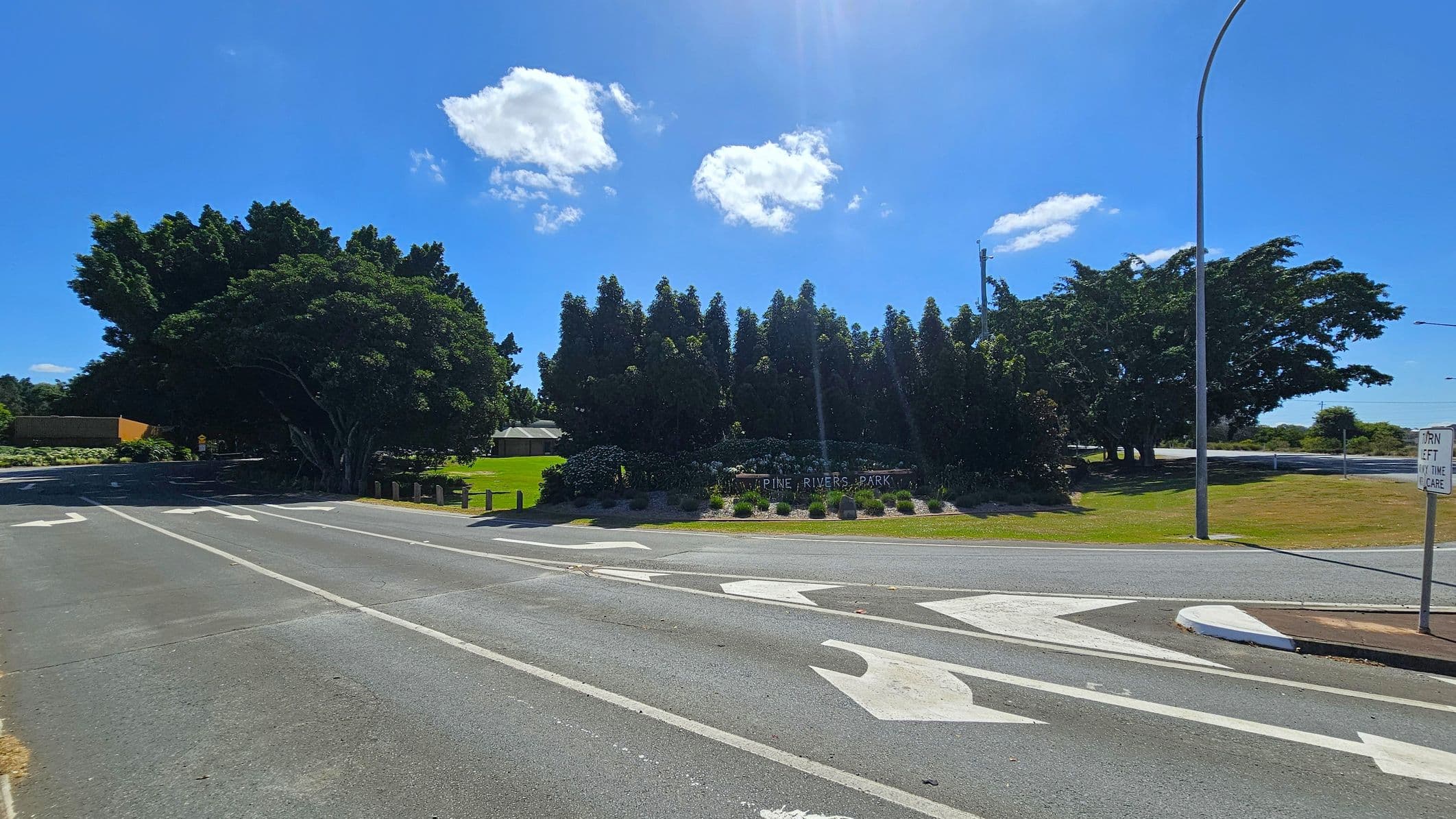 A road with white directional arrows painted on the surface. On the left, there are large, dense trees and a small building partially visible. In the centre, a sign reads "Pine Rivers Park" surrounded by neatly arranged bushes and flowers. On the right, a grassy area with a tall streetlight and a sign that says "Turn left any time with care." The sky is clear with a few clouds.