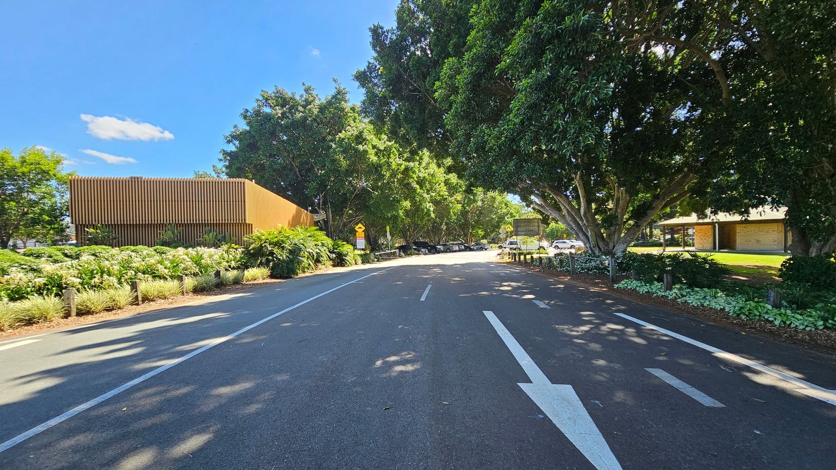 A road with a white arrow pointing forward runs through the centre. On the left, there's a modern building with vertical wooden slats, surrounded by lush greenery and plants. On the right, large trees provide shade, and a small building with a sloped roof is visible. Several cars are parked under the trees, and a signpost is near the road. The scene is well-lit, suggesting a sunny day.