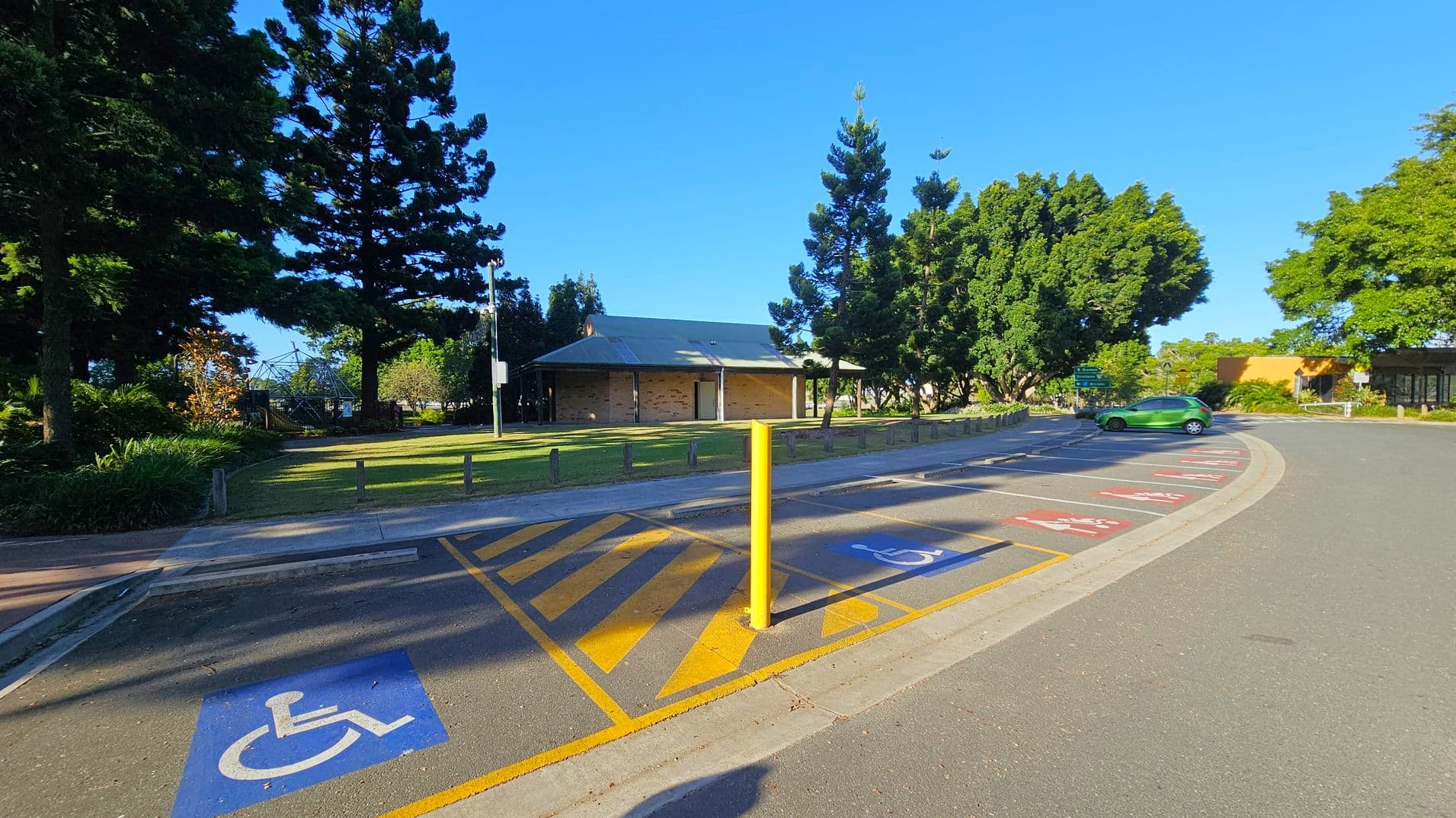 A parking area with designated disabled parking spaces marked in blue on the foreground. A yellow pole stands in the centre of the image. On the left, there are tall trees and a grassy area with a small building in the background. On the right, a green car is parked near more trees and a building with a yellow facade. The road curves around the parking area, and the sky is clear.