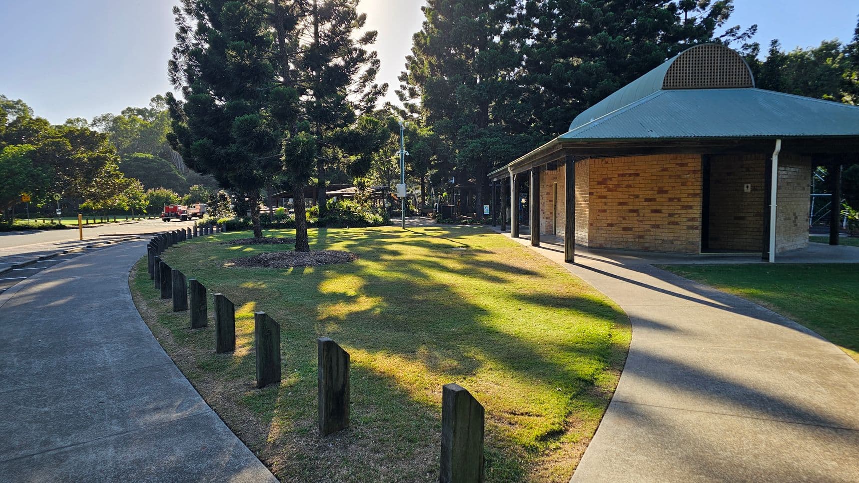 A curved concrete path runs through a grassy area, bordered by wooden posts on the left. Tall trees cast long shadows across the grass. On the right, a brick building with a metal roof stands, featuring a small porch area. In the background, more trees and a road with a red vehicle are visible. The scene is well-lit, suggesting a sunny day.