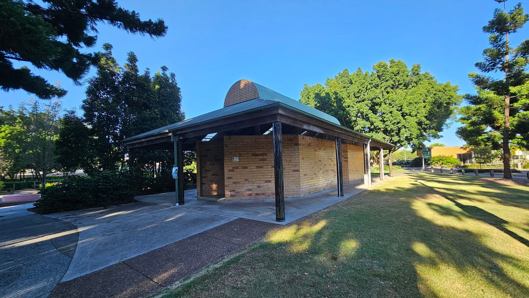 A small brick building with a green metal roof and a curved vent on top. It is supported by dark wooden posts. The building is surrounded by a paved area and grass. On the left, there are dense trees casting shadows, while on the right, more trees and a sign are visible. The sky is clear and blue.