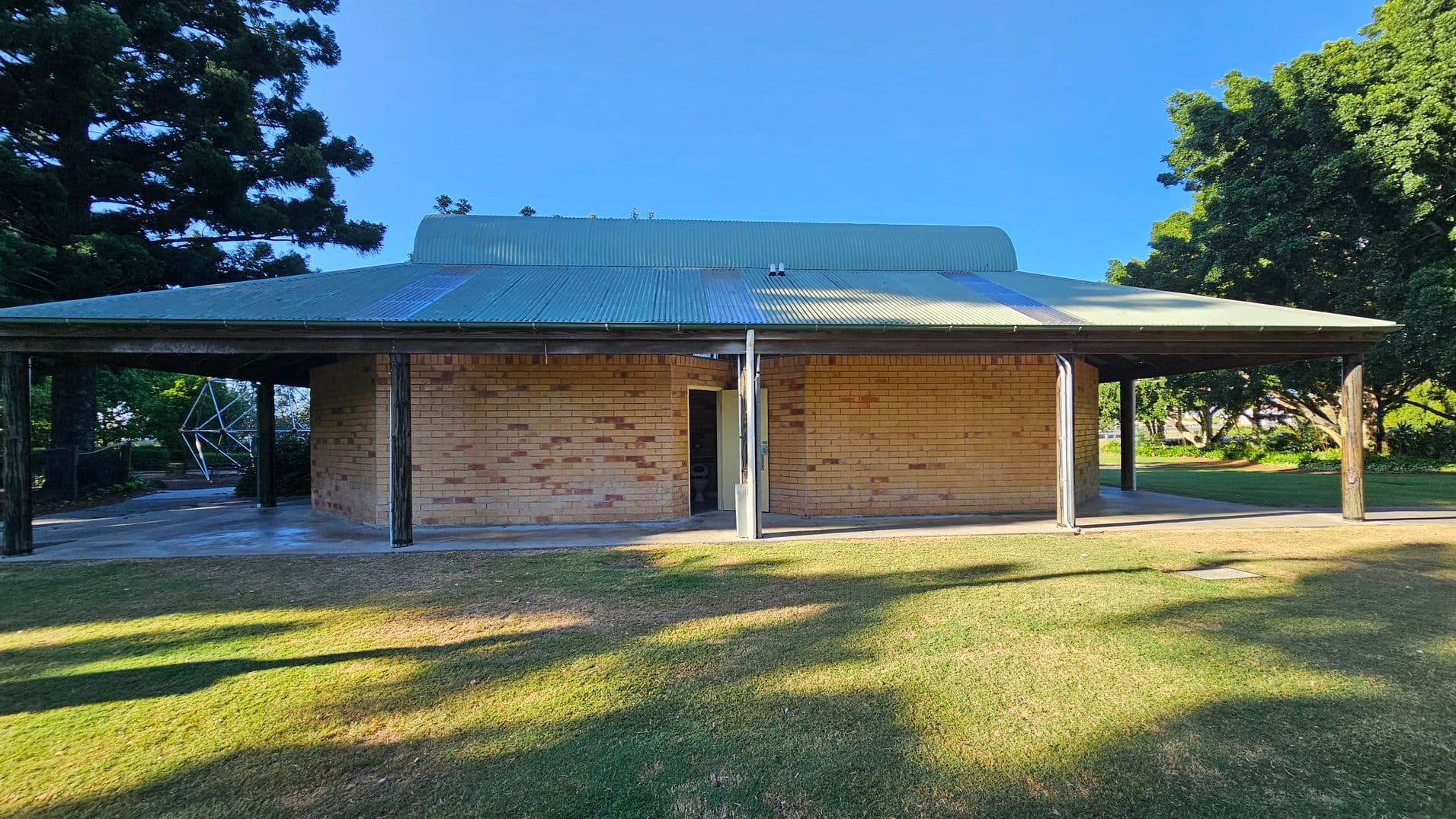 A brick building with a green corrugated metal roof is centred in the image. The structure has an open entrance and is supported by wooden posts. On the left, there is a large tree casting shadows on the grass. To the right, more trees are visible, providing shade. The foreground features a grassy area with patches of sunlight and shadow. The background includes additional greenery and a glimpse of a geometric metal structure.