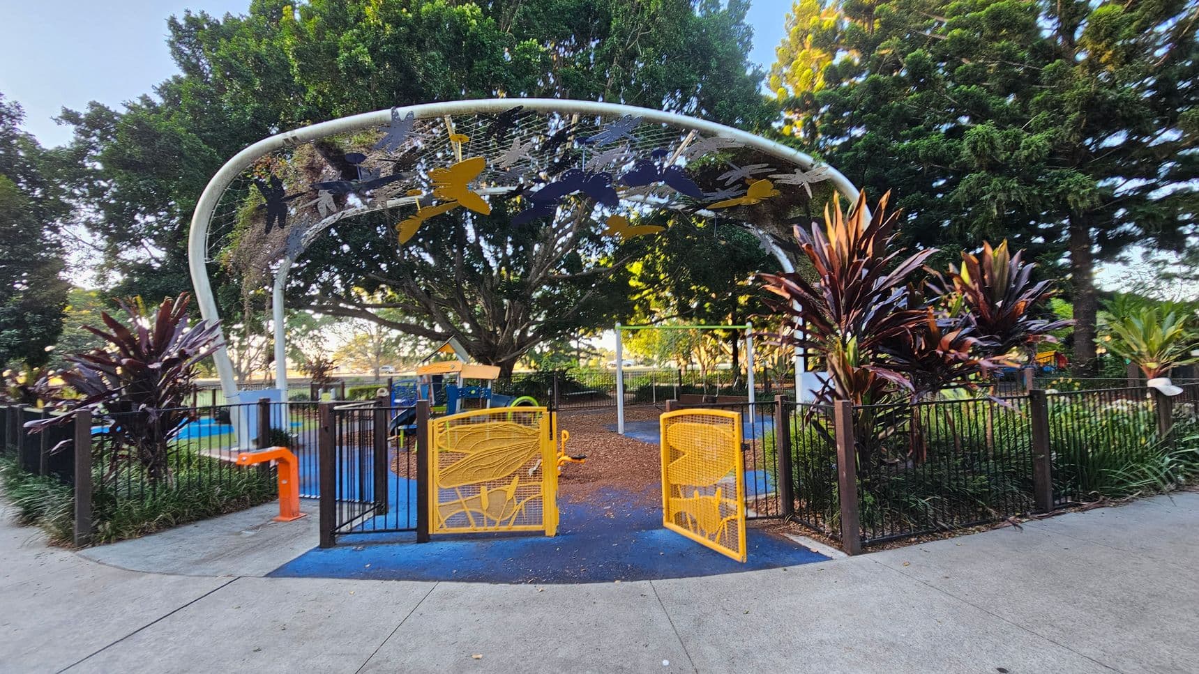 A playground entrance with bright yellow gates featuring leaf designs. The gates are set within a black metal fence. Above, a white archway is adorned with colourful butterfly and leaf decorations. The ground is a mix of concrete and blue safety flooring. Lush greenery, including large trees and red-leaved plants, surrounds the area. In the background, playground equipment is visible under the shade of a large tree.