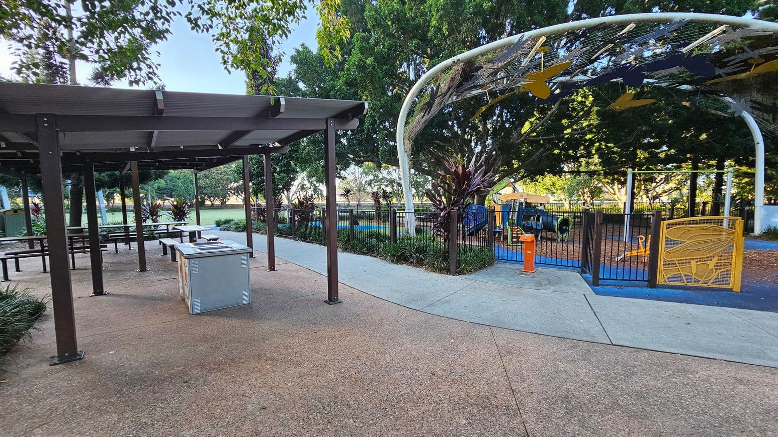 A park scene with a covered picnic area on the left, featuring several tables and a barbecue grill on a speckled concrete floor. To the right, a fenced playground with a colourful play structure is visible, surrounded by trees and greenery. The playground entrance has a yellow gate, and a decorative arch with hanging elements is overhead. A pathway runs between the picnic area and the playground, with trees and plants in the background.
