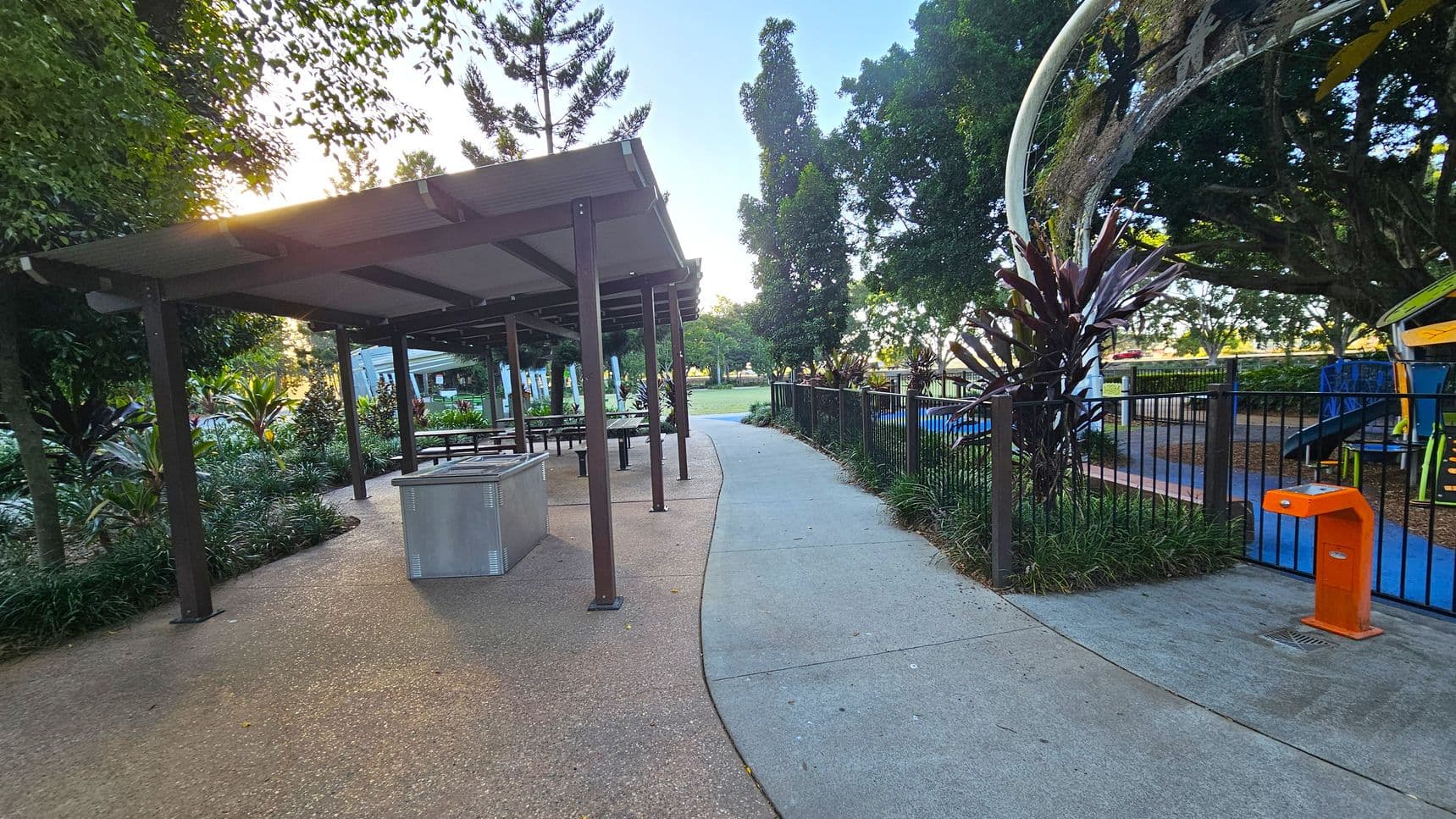A park scene with a covered picnic area on the left, featuring tables and benches under a metal roof. The ground is a mix of concrete and gravel. On the right, a fenced playground with colourful equipment is visible, surrounded by trees and plants. A curved concrete path runs through the middle, leading into the distance. An orange water fountain is near the playground fence. Tall trees and lush greenery are in the background.