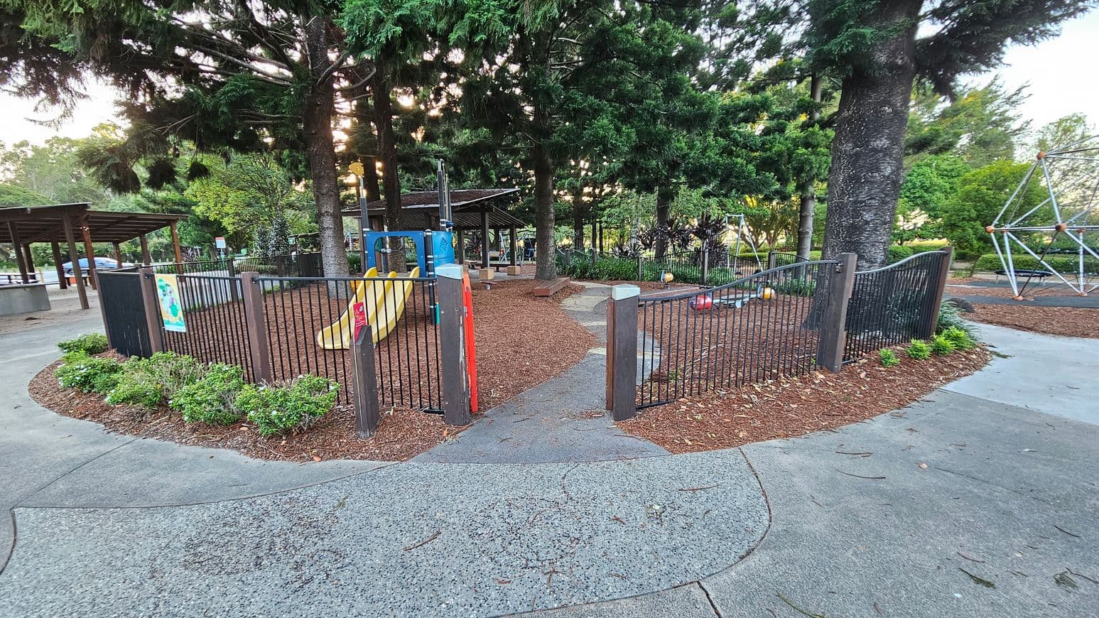 A playground surrounded by a black metal fence with a gate in the centre. Inside, there is a yellow slide and various play equipment. The ground is covered with wood chips. Tall trees provide shade, and there is a path leading to the entrance. On the left, a covered area with a roof is visible, and on the right, a geometric climbing structure. The foreground has a concrete path with some scattered leaves.