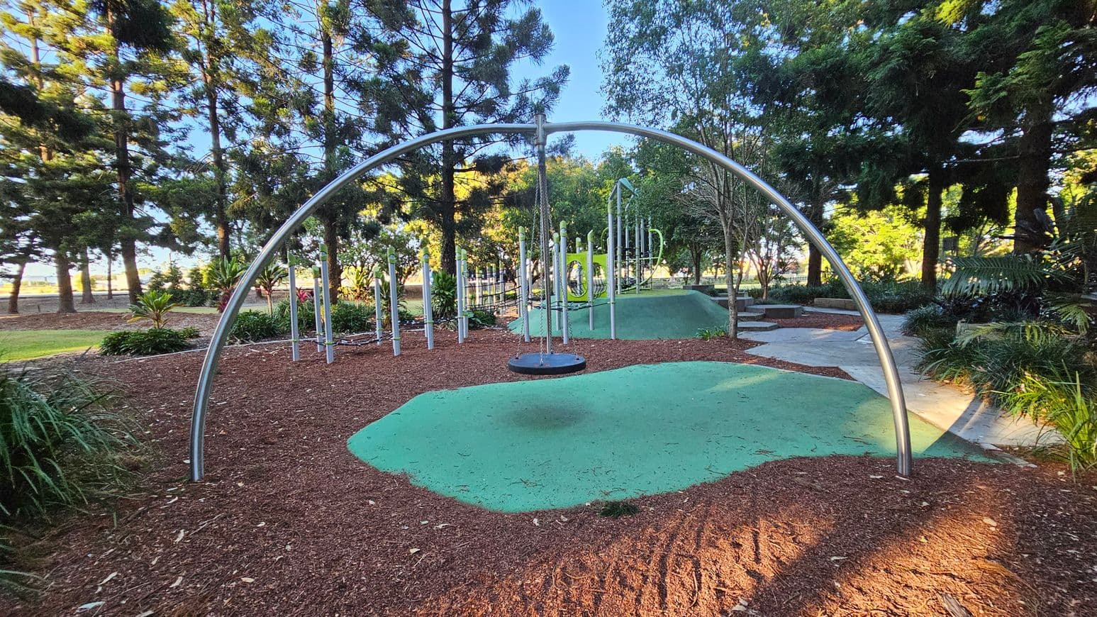 A playground with a large metal arch structure in the centre, supporting a circular swing. The ground is covered with a mix of green safety surfacing and brown mulch. In the background, there are various climbing structures and poles. Tall trees and lush greenery surround the area, providing shade and a natural setting. The path on the right is paved, leading through the park.