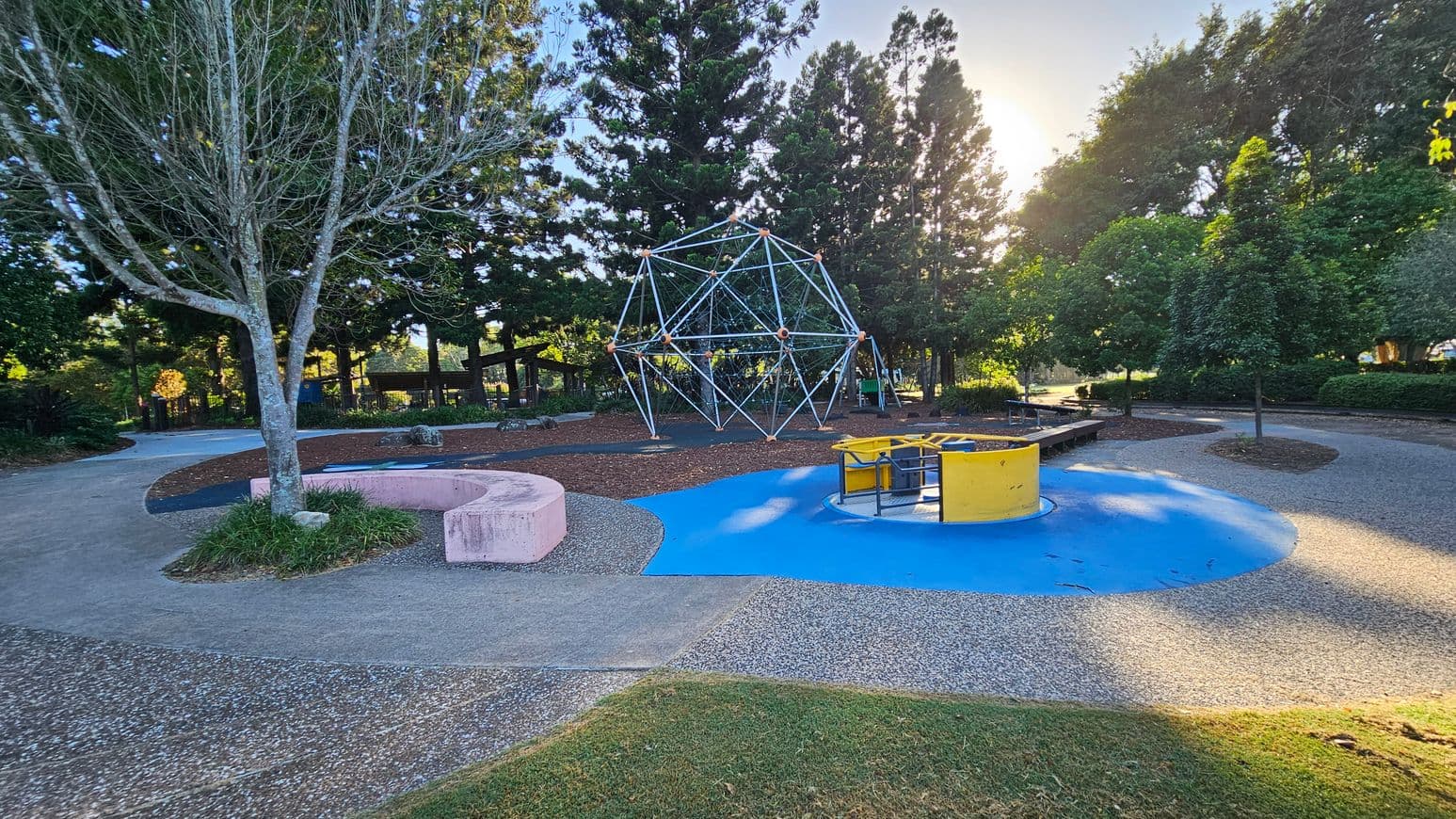 A playground with a geometric climbing structure in the centre, surrounded by tall trees. On the left, a bare tree stands next to a curved pink concrete bench. On the right, a yellow and blue roundabout sits on a blue rubber surface. The ground is a mix of grass, concrete, and pebbled paths. The background is filled with dense greenery and more trees, with sunlight filtering through.