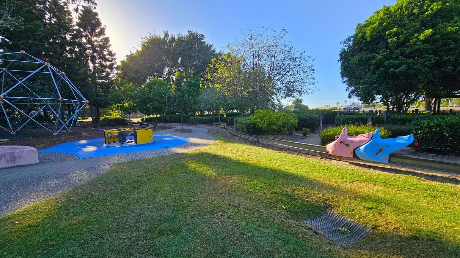 A playground with a grassy foreground and a path leading through it. On the left, there's a large geometric climbing structure made of metal bars. In the centre, a roundabout with yellow and black panels sits on a blue surface. On the right, two animal-shaped spring riders, one pink and one blue, are positioned side by side. The background is filled with trees and bushes, providing shade and greenery.
