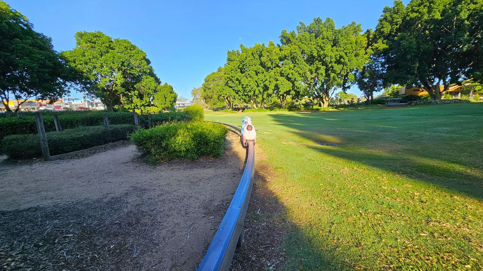 A park scene with a narrow metal track running through the centre. A small, toy-like train is positioned on the track. On the left, there is a dirt path bordered by low hedges and trees. On the right, a grassy area extends with large trees providing shade. The background features more trees and a glimpse of buildings. The ground is a mix of grass and dirt, and the area is well-lit by sunlight.