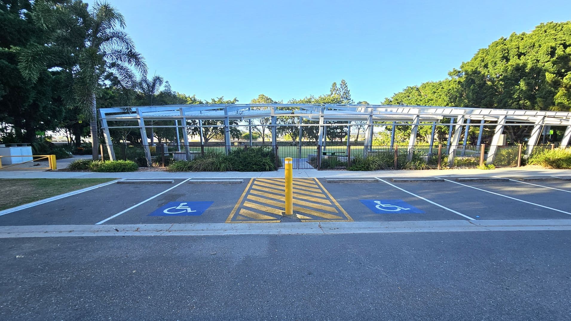 A parking area with two marked disabled parking spaces on either side of a yellow-striped zone with a yellow bollard in the centre. In the background, there is a metal structure with a roof, surrounded by greenery and trees. The ground is paved, and the area is well-maintained with some grass visible near the parking spaces.