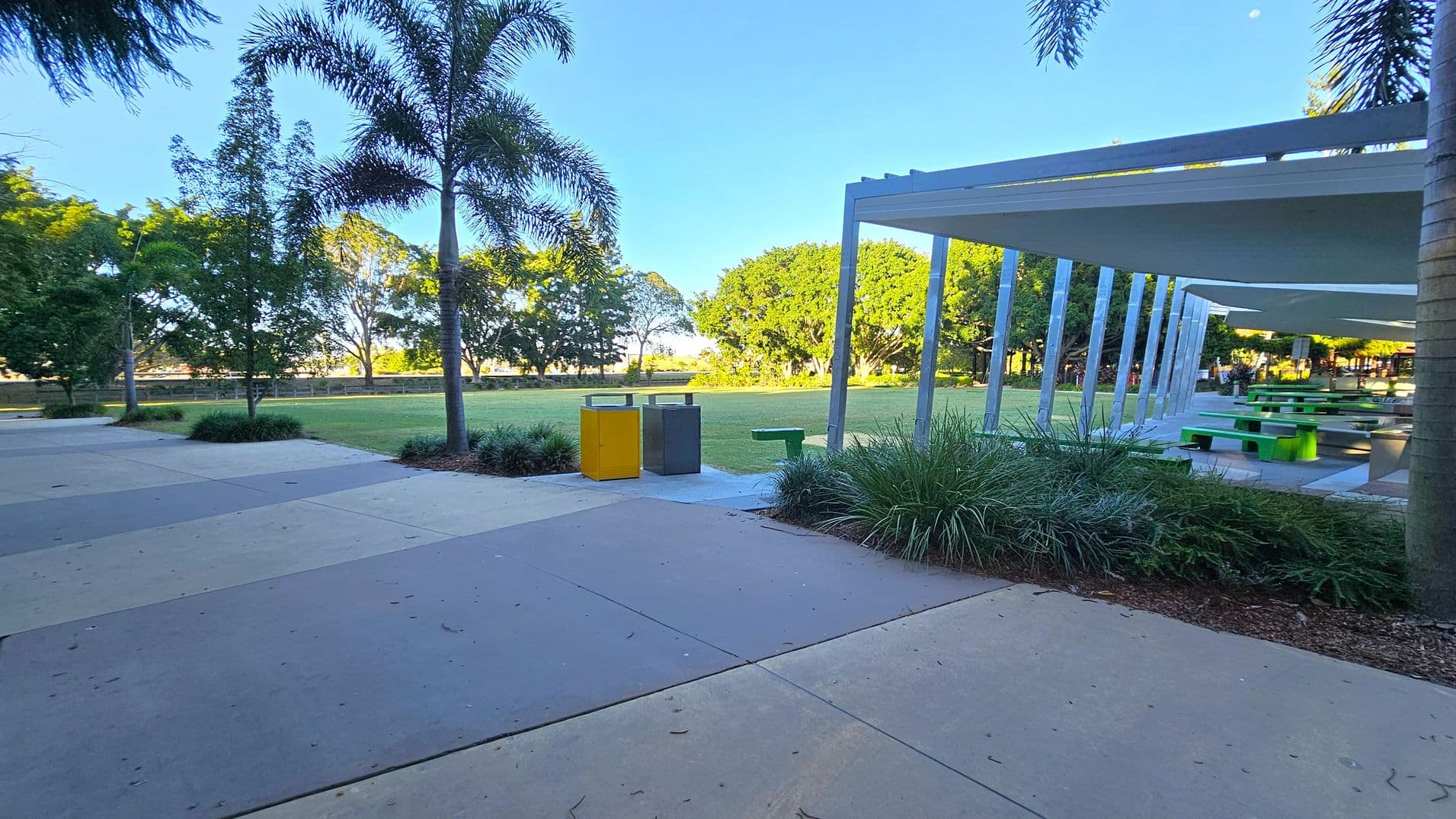 A park scene with a paved area in the foreground. On the left, there are palm trees and a couple of rubbish bins, one yellow and one grey. In the middle, a grassy field is bordered by more trees. On the right, a covered seating area with green picnic tables is visible, supported by white beams. The background features more trees and a clear sky.