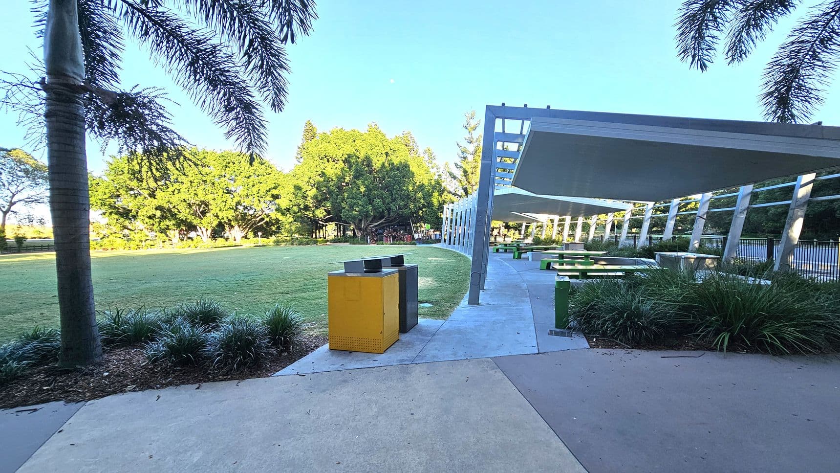 A park scene with a grassy area on the left, bordered by trees. In the foreground, a palm tree and a pair of yellow and black bins are visible. On the right, a covered area with a modern metal structure provides shade over green picnic tables. The ground is a mix of concrete and grass, with landscaped plants along the edges. The ceiling of the structure is flat and extends over the seating area.