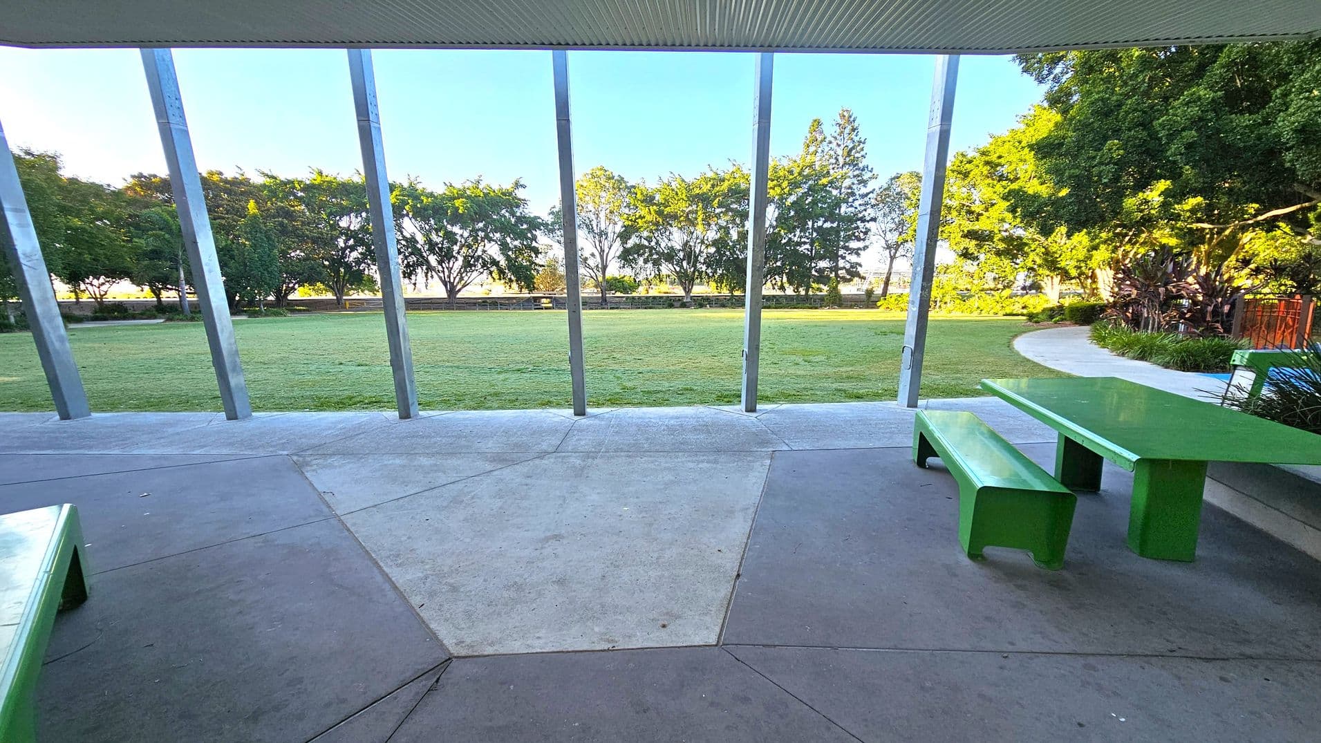 A covered outdoor area with a green picnic table and bench on the right. The floor is made of concrete with geometric patterns. In the background, a grassy field is bordered by a row of trees. Metal beams support the structure, and a pathway curves around the right side, lined with plants. The ceiling is corrugated.