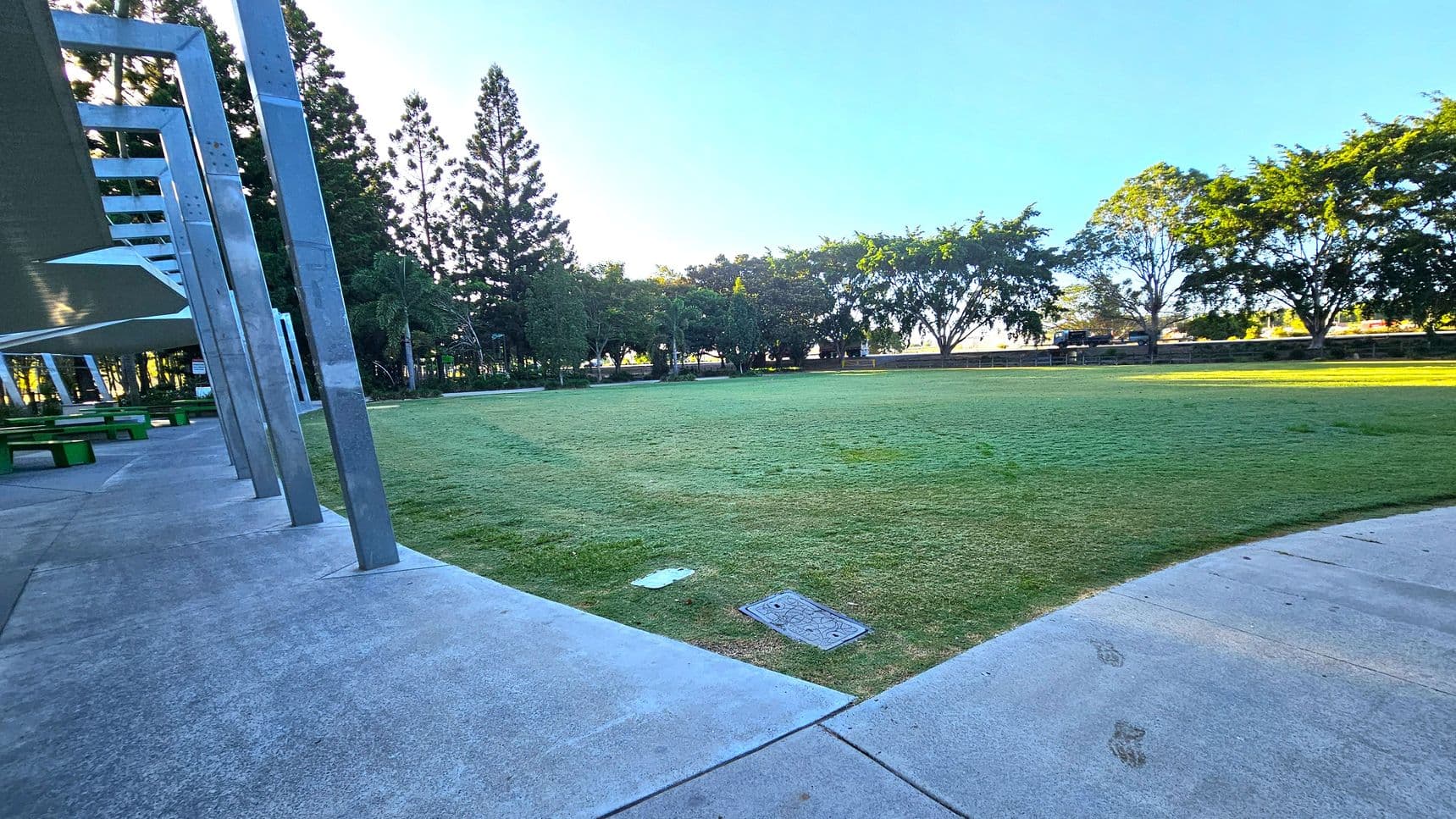 A grassy park area with a concrete path curving through the foreground. On the left, there are metal beams and a covered area with green benches underneath. Tall trees line the background, providing shade and a natural border. The sky is clear, and the area is well-lit, suggesting daytime.