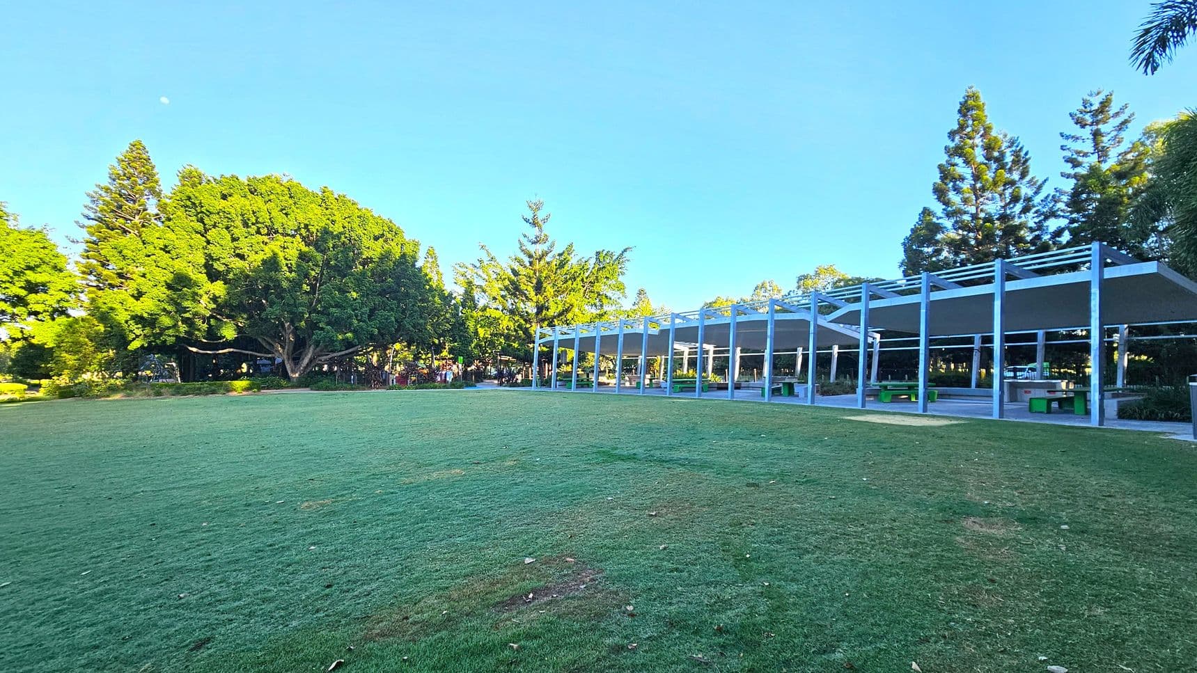 A grassy field stretches across the foreground, with a large, leafy tree and several tall coniferous trees on the left. On the right, a modern structure with a series of open, metal-framed shelters provides shaded seating areas. The background is filled with more trees, creating a lush, green environment. The sky is clear and blue.