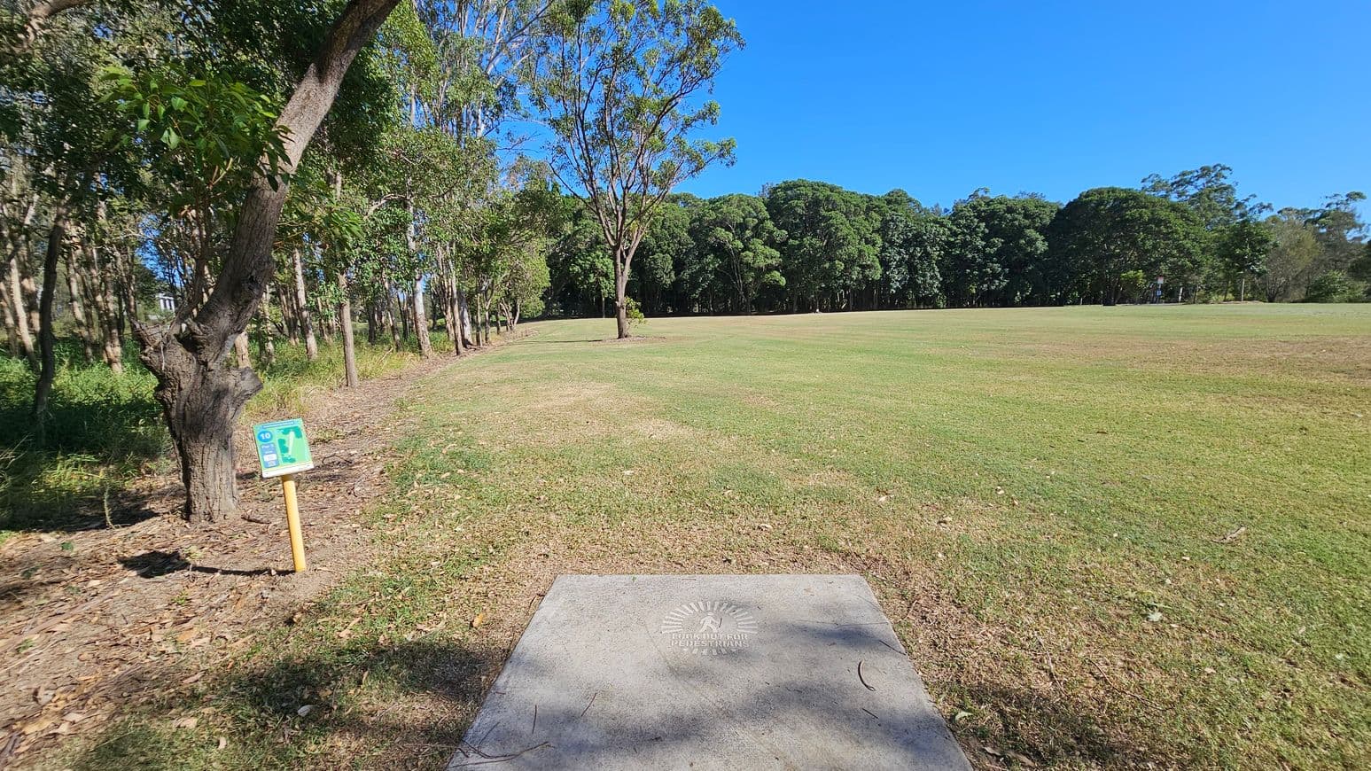 A grassy field with a concrete slab in the foreground, marked with a warning for pedestrians. To the left, a row of trees lines the edge of the field, with a small signpost displaying a map or instructions. In the background, a dense cluster of trees forms a natural boundary. The ground is a mix of green grass and patches of dry, brown grass.